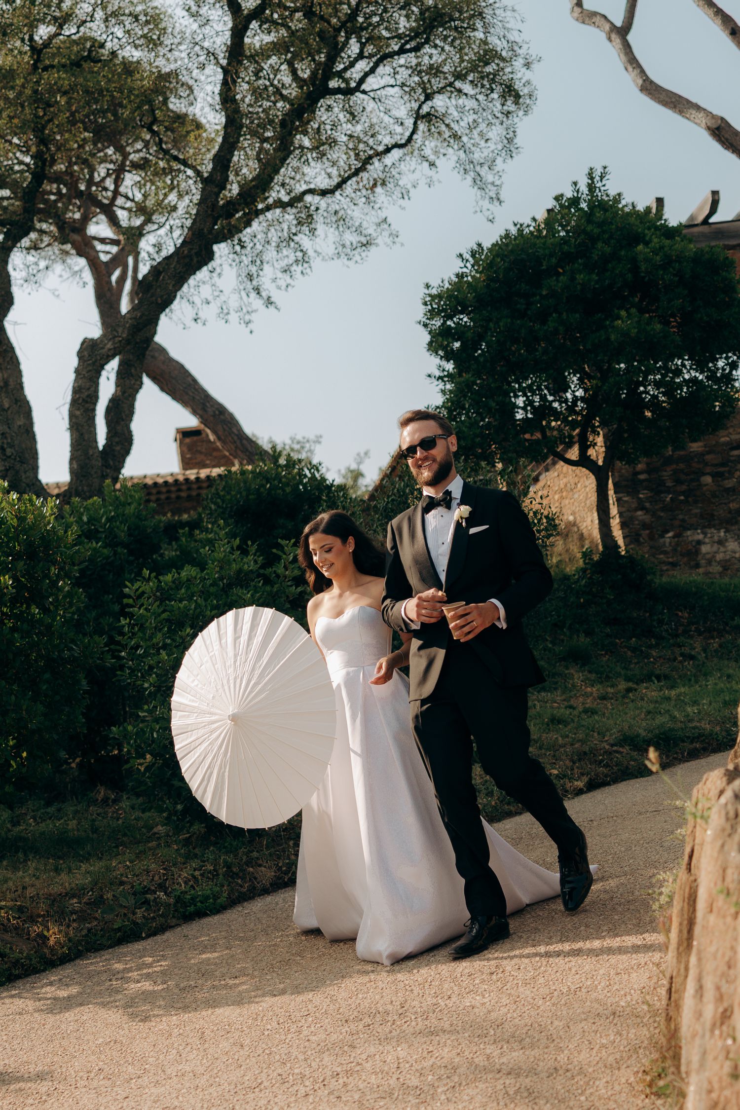 A wedding couple poses with a white parasol in a garden setting with trees and rustic architecture.