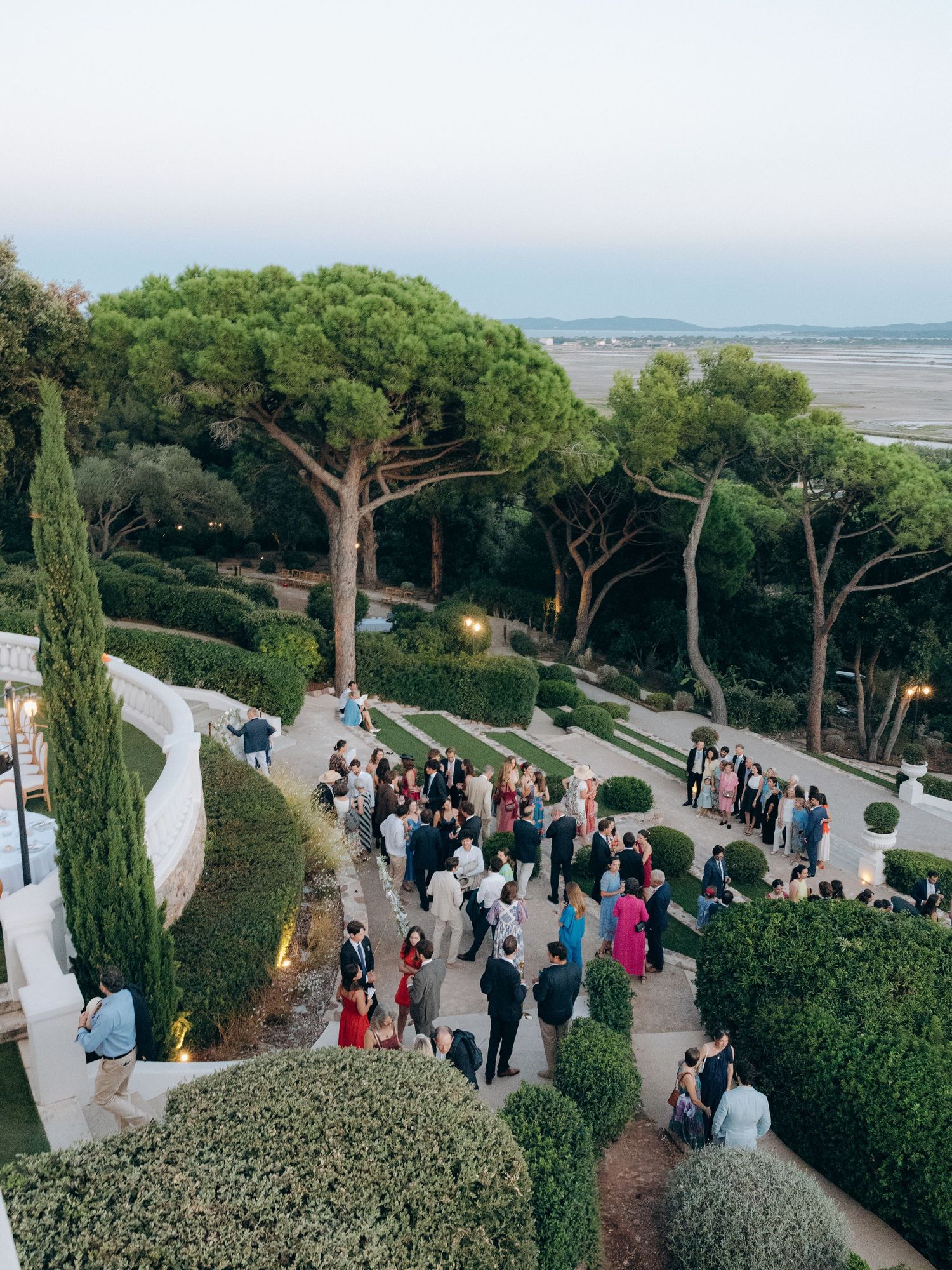 Aerial view of an elegant outdoor wedding reception on terraced gardens with lush greenery and scenic coastal views at dusk.