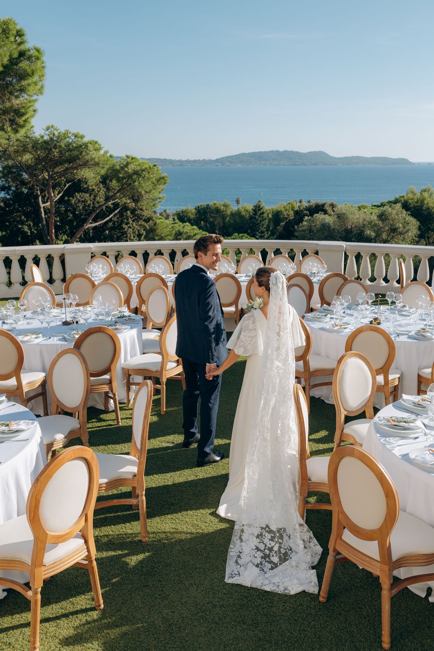 Bride and groom walk among elegant round tables set for outdoor wedding reception with ocean views.