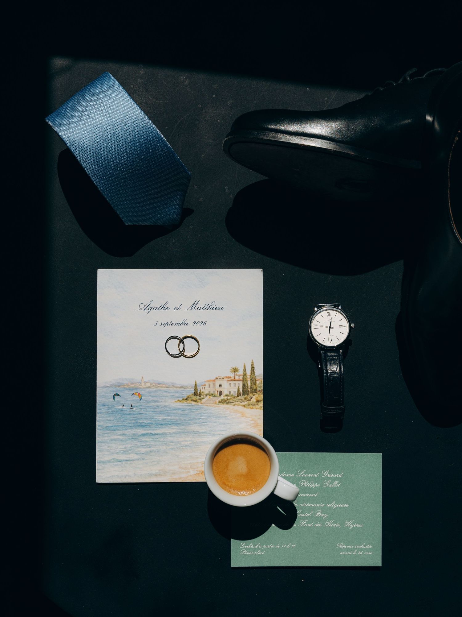 Elegant wedding flat lay with invitation, espresso, watch, blue tie, and black dress shoes on dark surface.