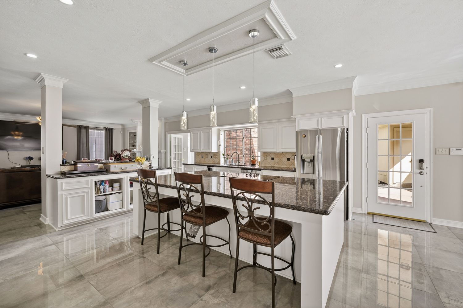 Elegant white kitchen with dark granite island, bar stools, marble floors, and stainless steel appliances in a luxury home.