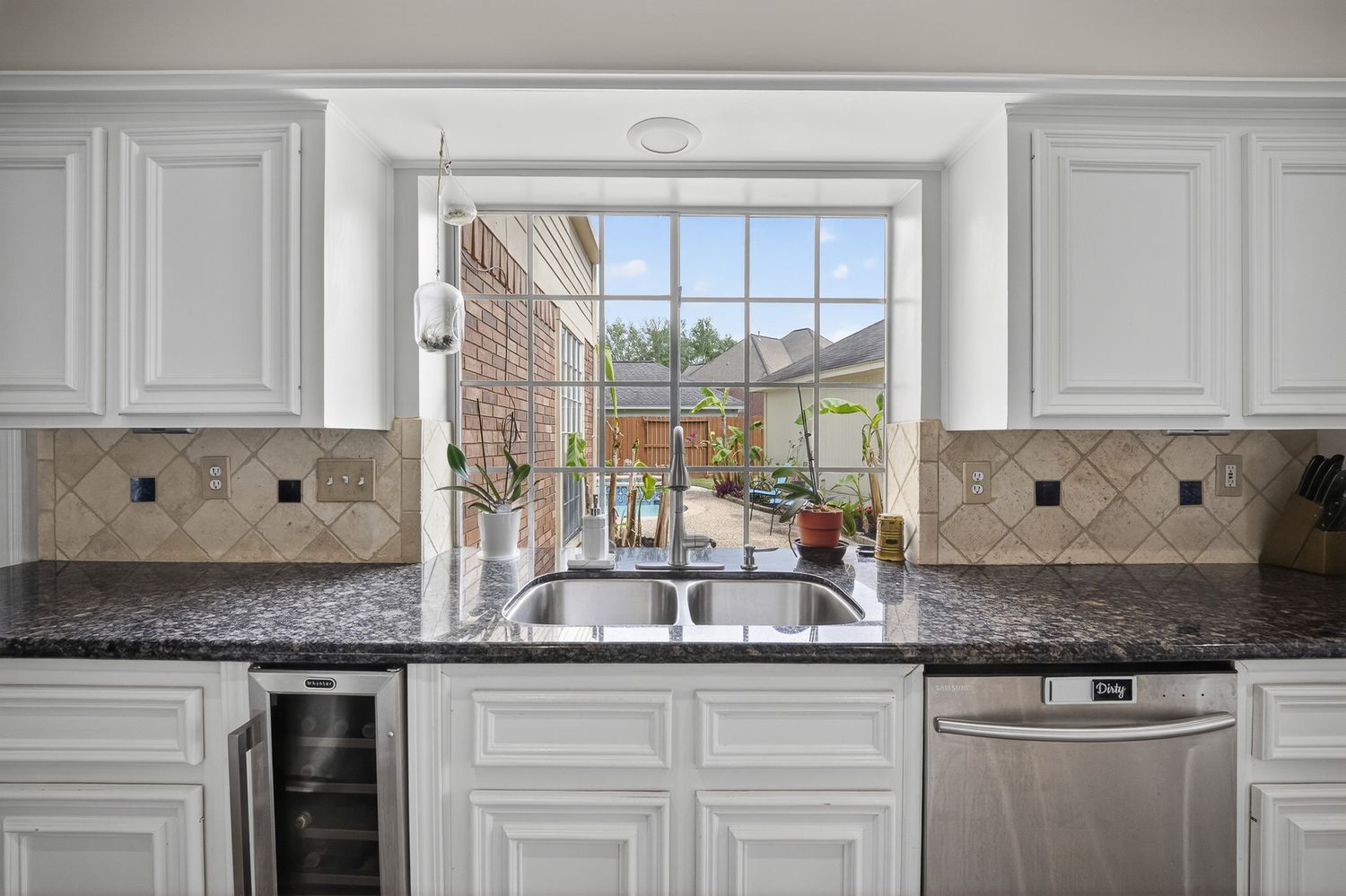 White kitchen cabinets with dark granite countertops, tile backsplash, double sink, and a bright garden window overlooking a patio.