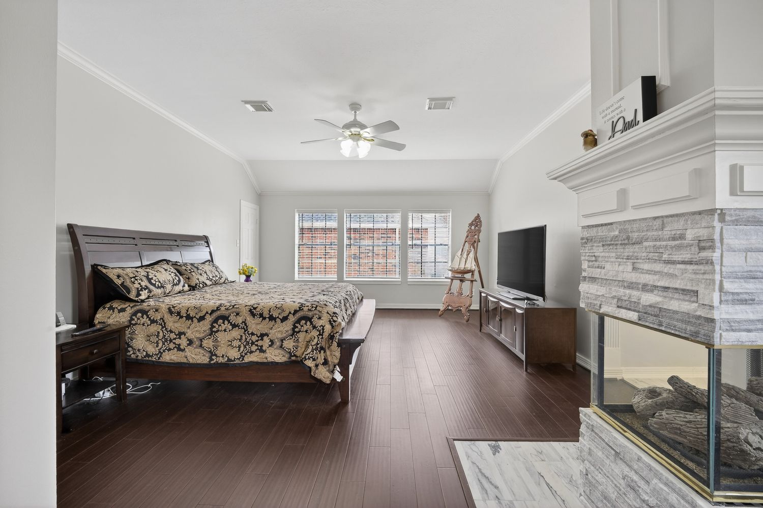 Spacious master bedroom with dark hardwood floors, ceiling fan, stone fireplace, and large windows providing natural light.