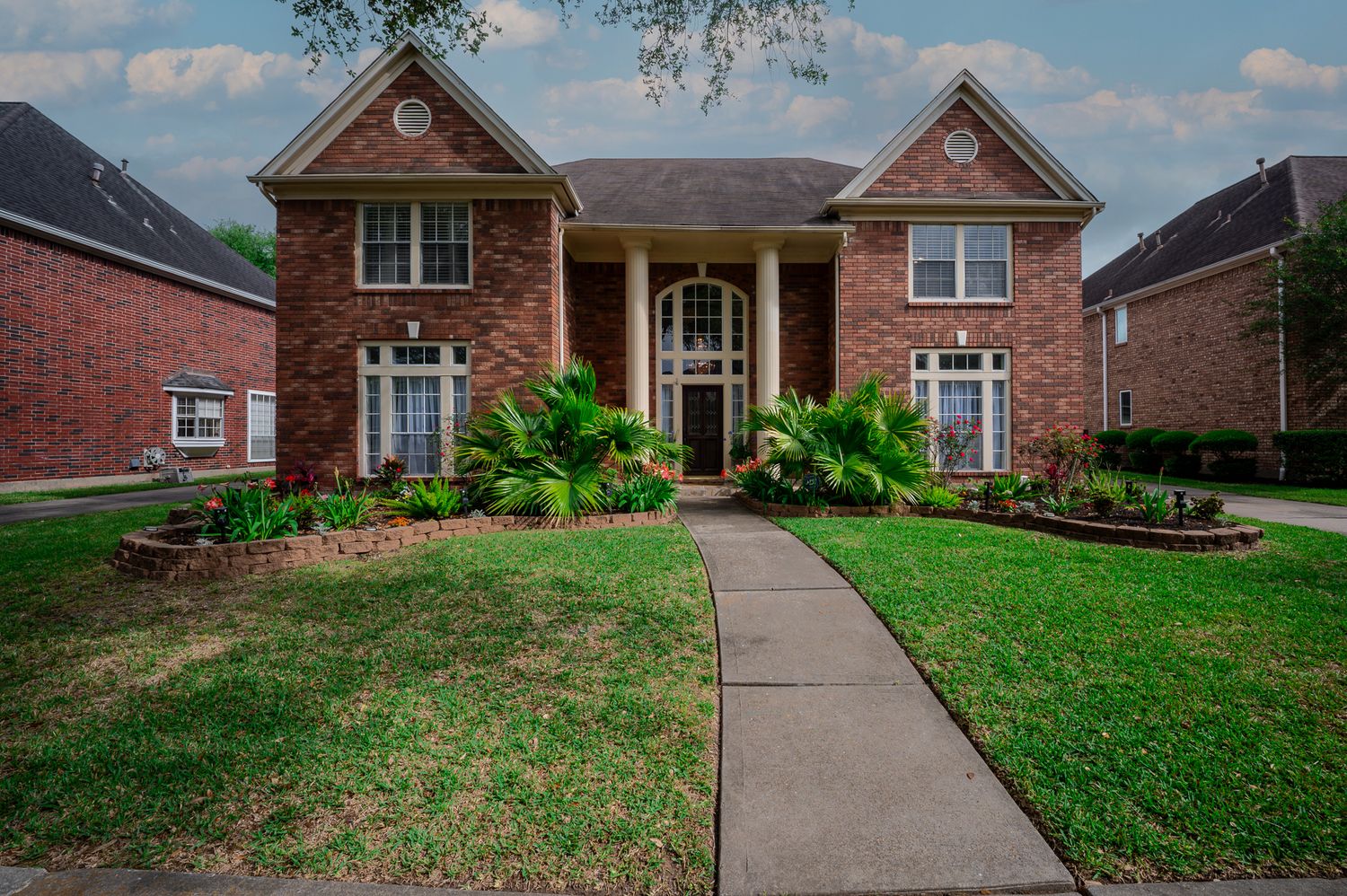 Large two-story brick home with manicured lawn, lush landscaping, and a concrete walkway leading to an arched front entry.