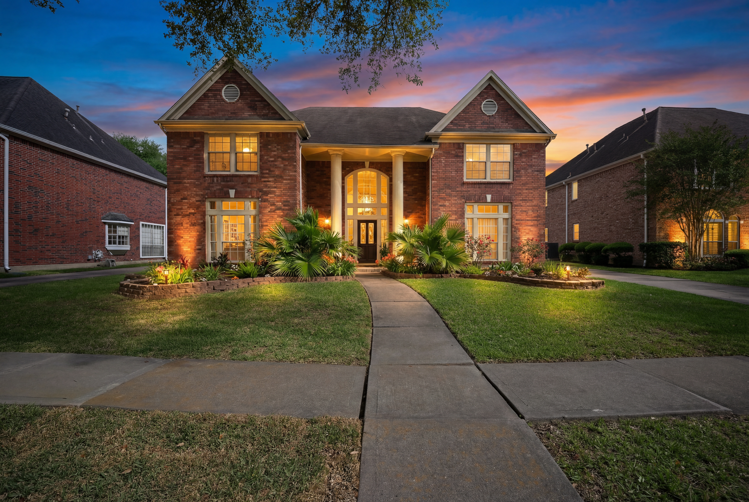 Two-story brick home with elegant columns, lush landscaping, and warm exterior lighting at dusk.