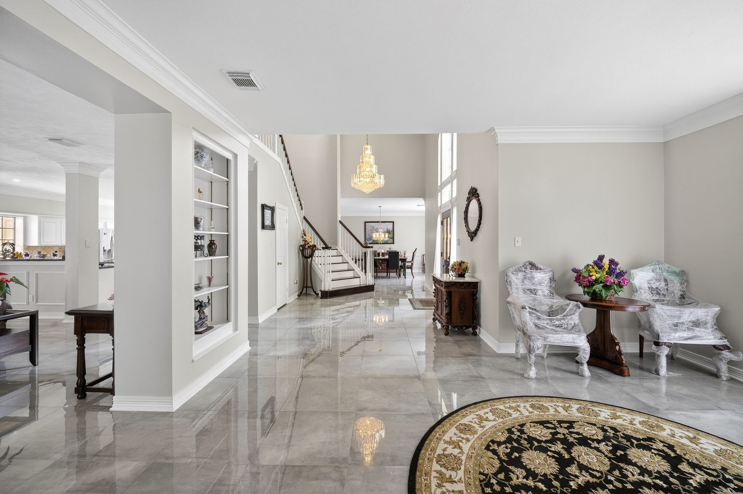 Elegant foyer with polished marble floors, white columns, grand staircase, and chandelier in a luxurious home.