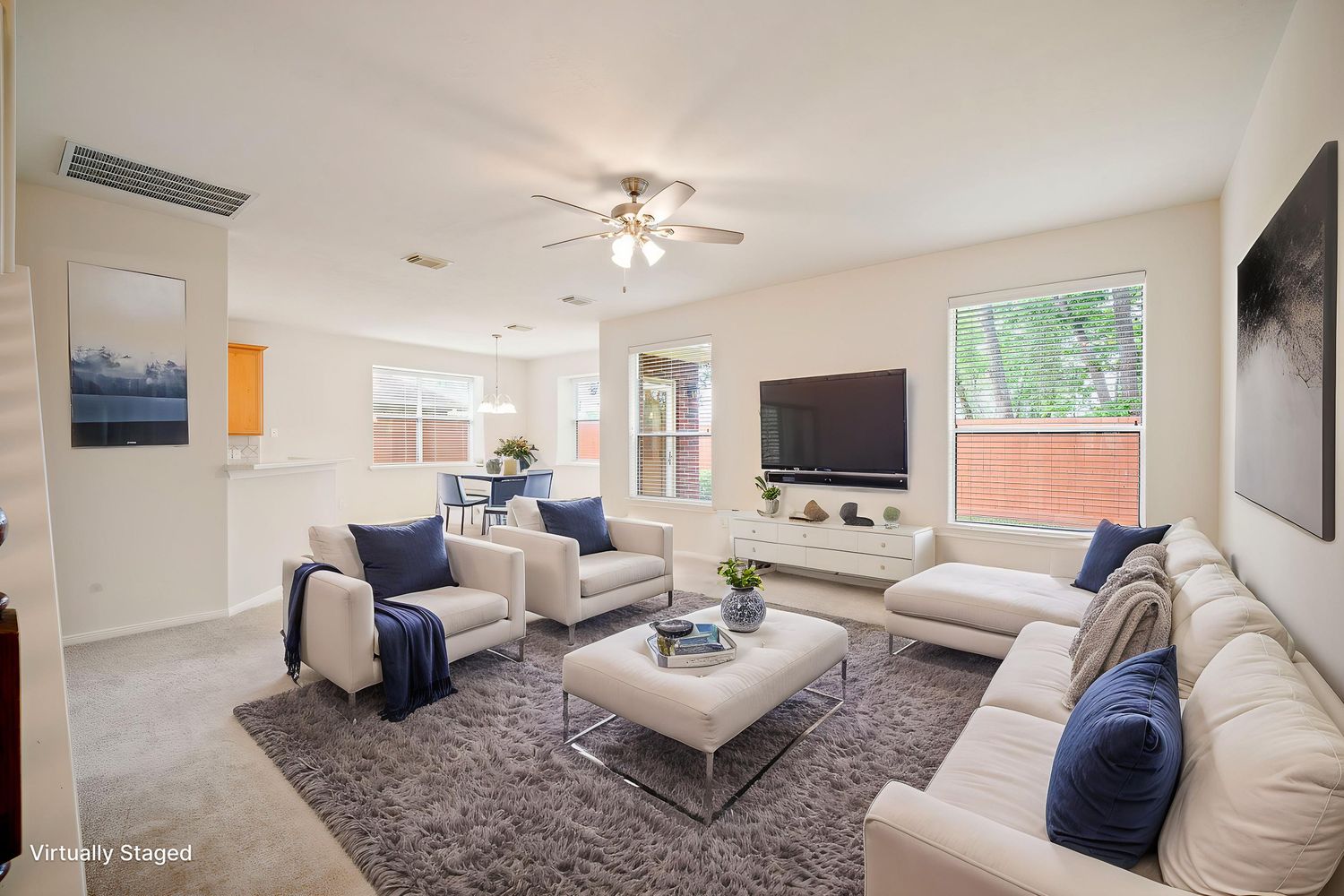 Bright living room with white sofas, navy blue accent pillows, ceiling fan, large TV, and natural light from multiple windows.