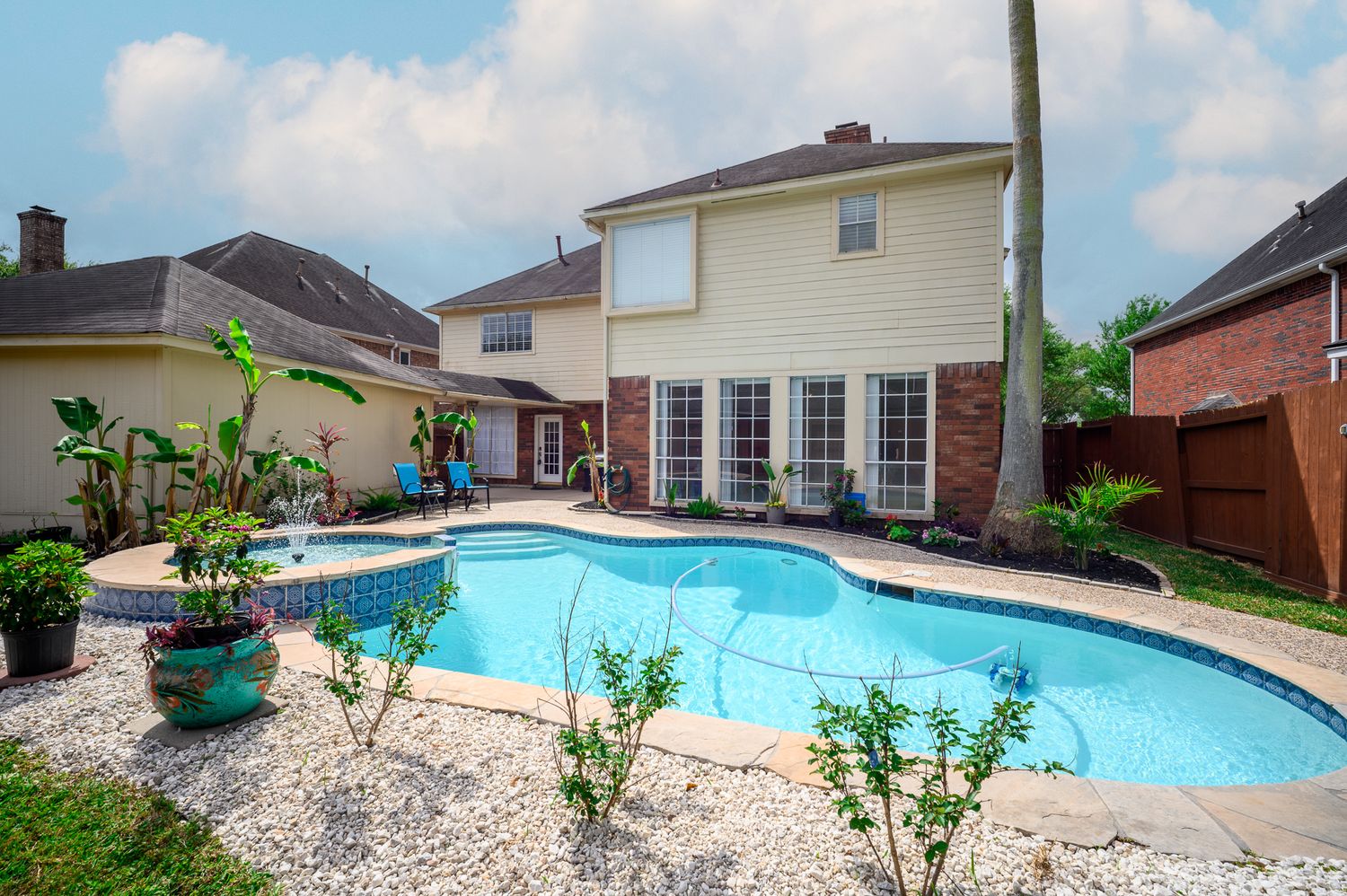 Backyard oasis featuring a large freeform swimming pool with fountain, patio seating, and lush landscaping behind a two-story home.