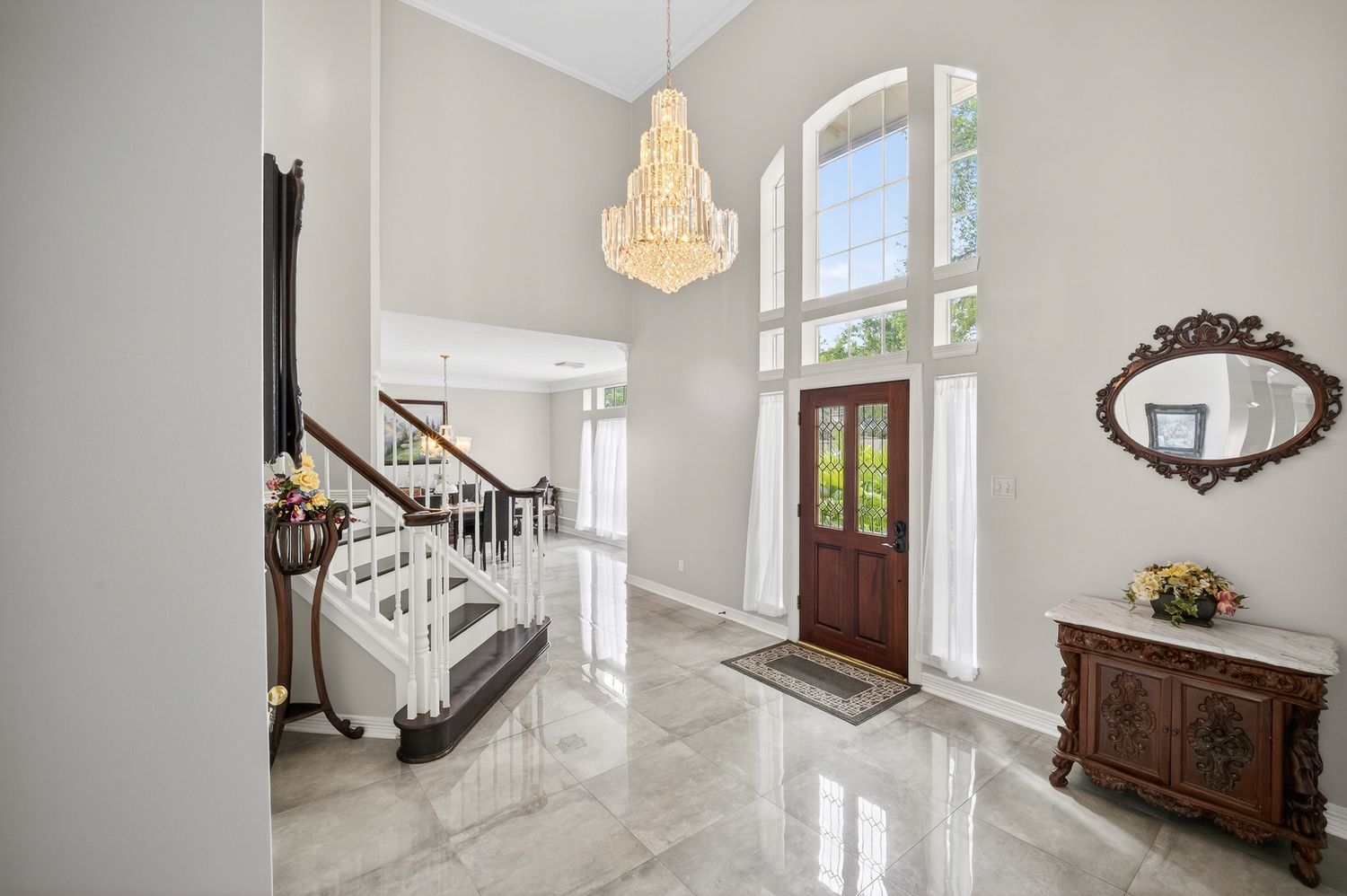 Elegant foyer with marble floors, grand chandelier, sweeping staircase, and arched wooden front door with tall windows.
