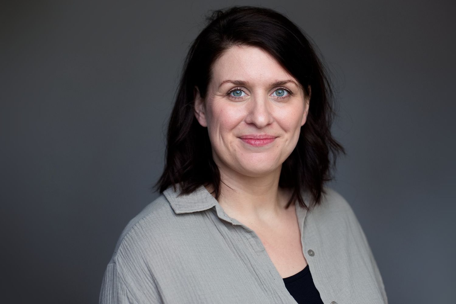 Portrait einer Frau in einer offenen grüngrauen Bluse passend zu ihrer Augenfarbe und einem schwarzen T-Shirt darunter.