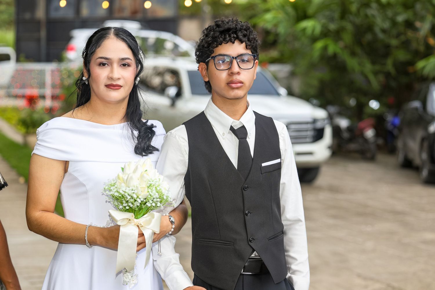 Young couple in formal wedding attire walk together outdoors, bride holding white flower bouquet.