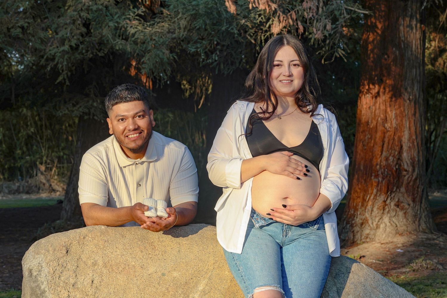 Expectant couple poses outdoors on a large rock surrounded by trees.