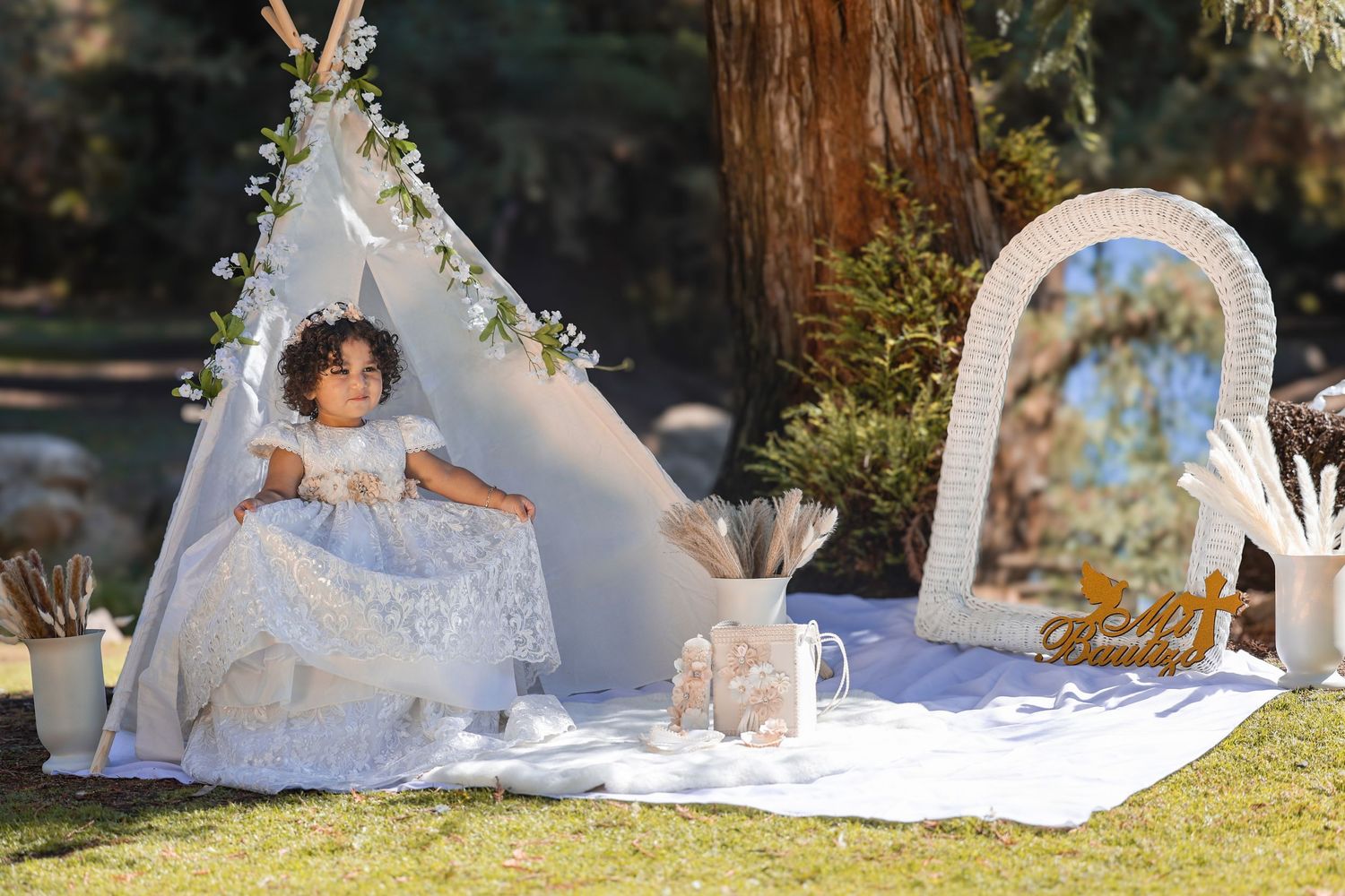 A toddler girl wearing an elegant white lace gown poses inside a floral-adorned teepee outdoors.