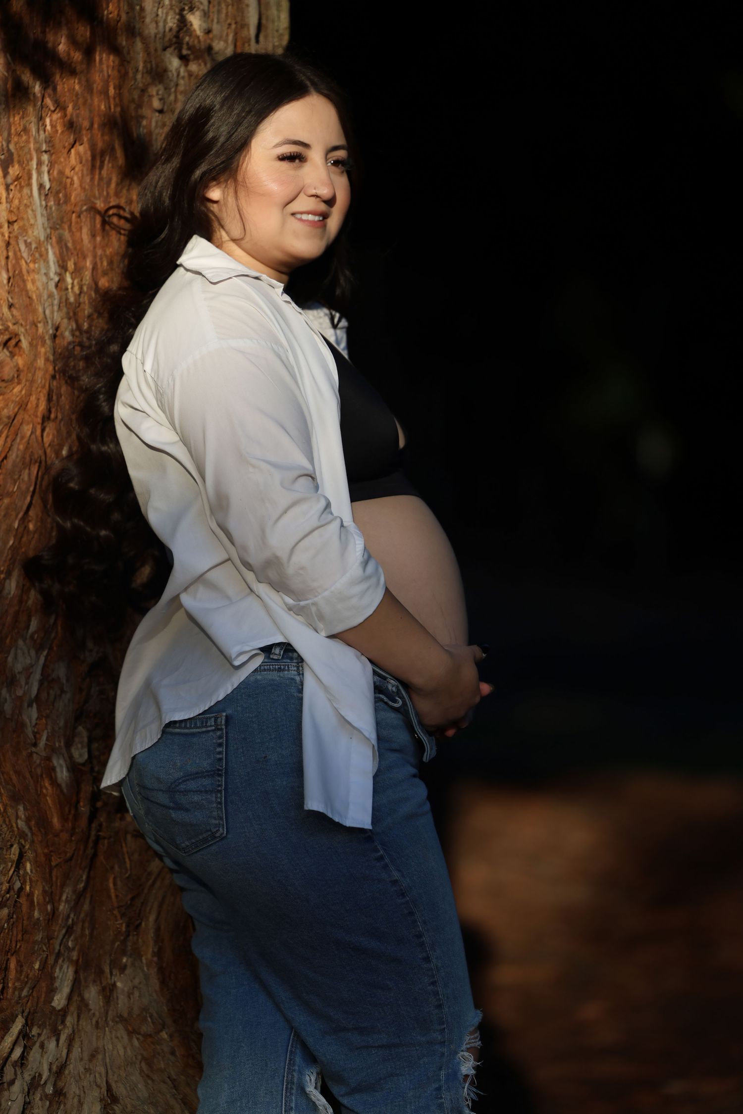 Smiling pregnant woman in white shirt and jeans cradles baby bump beside a tree outdoors.