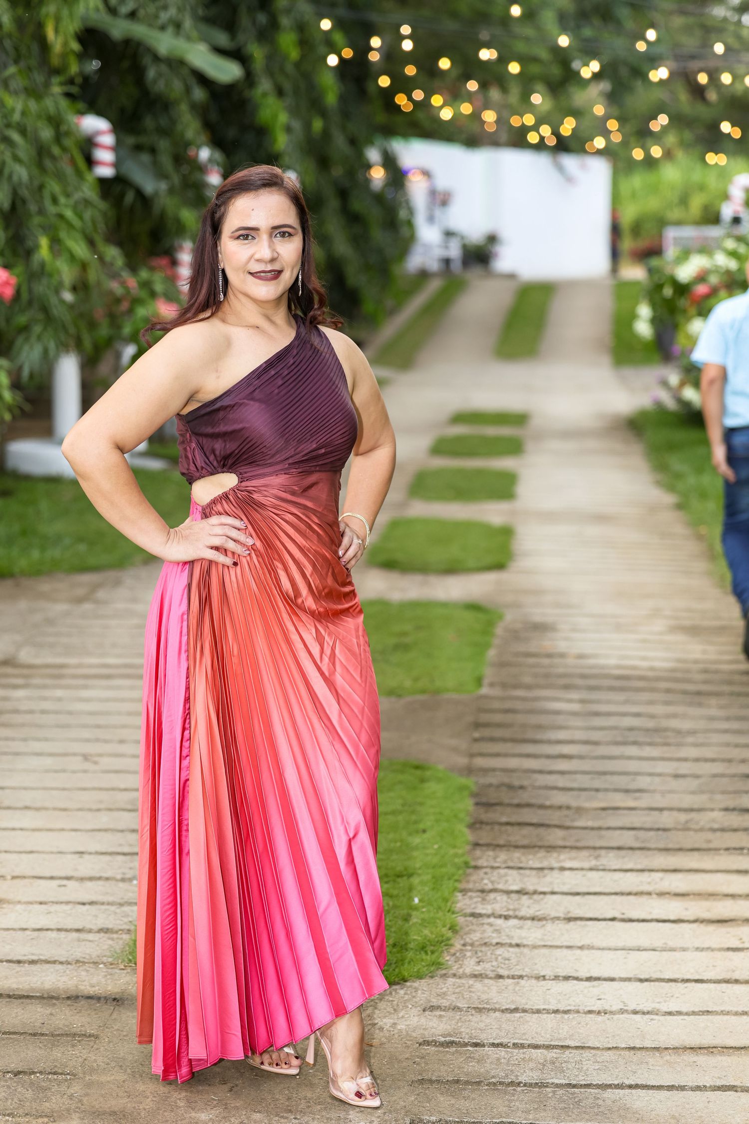 Woman in elegant ombre pink-purple one-shoulder pleated gown posing at outdoor garden event.
