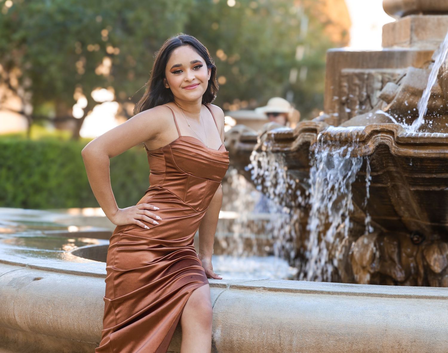 Young woman in copper satin gown poses elegantly beside ornate stone fountain at outdoor venue.