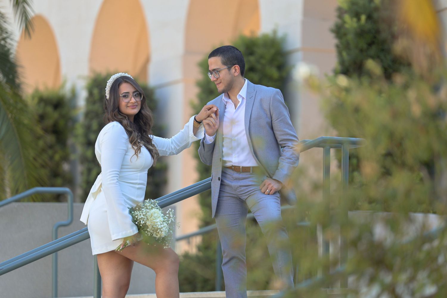 Bride in white mini dress and groom in gray suit share romantic moment on outdoor steps.