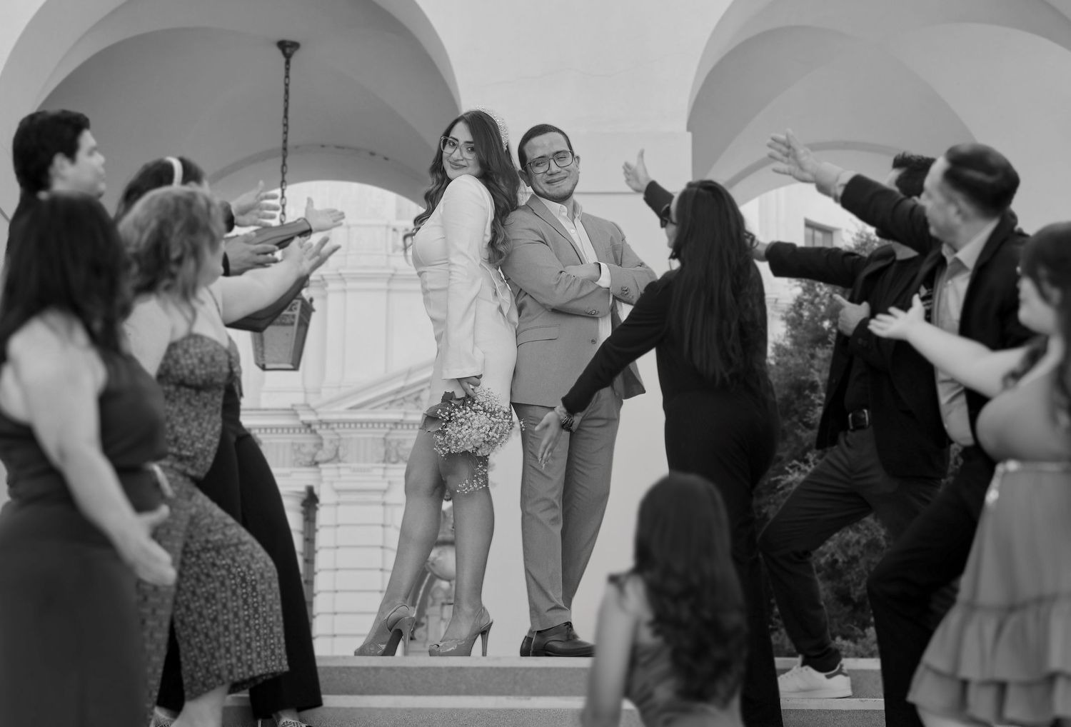 Black and white photo of couple standing on steps surrounded by cheering wedding guests outdoors.
