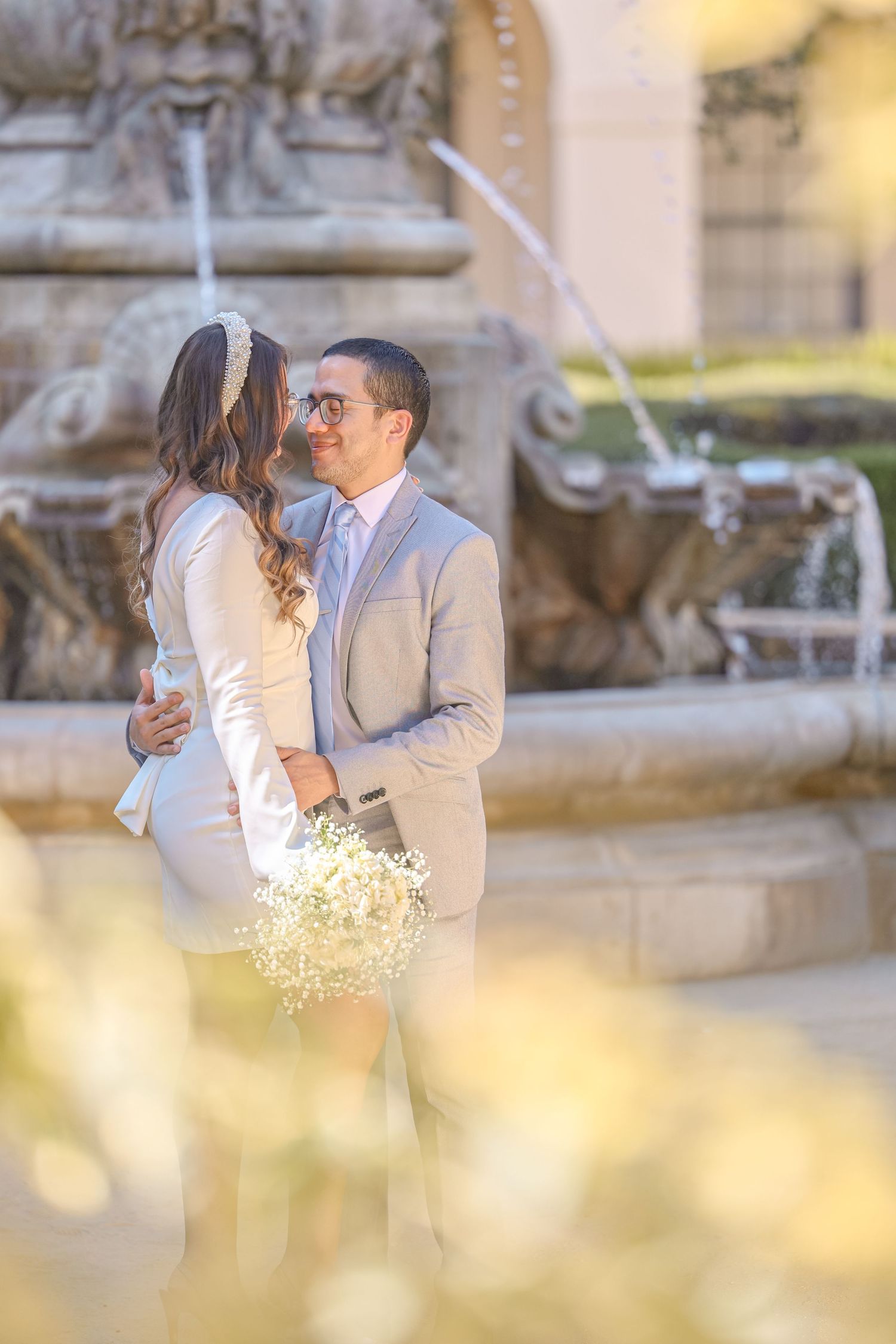 Couple embraces romantically in front of ornate stone fountain, groom in tan suit holding bouquet.