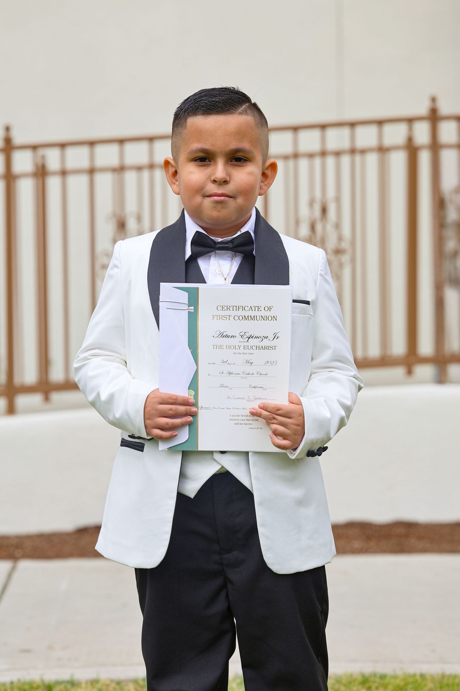 Young boy in white tuxedo holds First Communion certificate, dressed formally with bow tie for religious milestone.