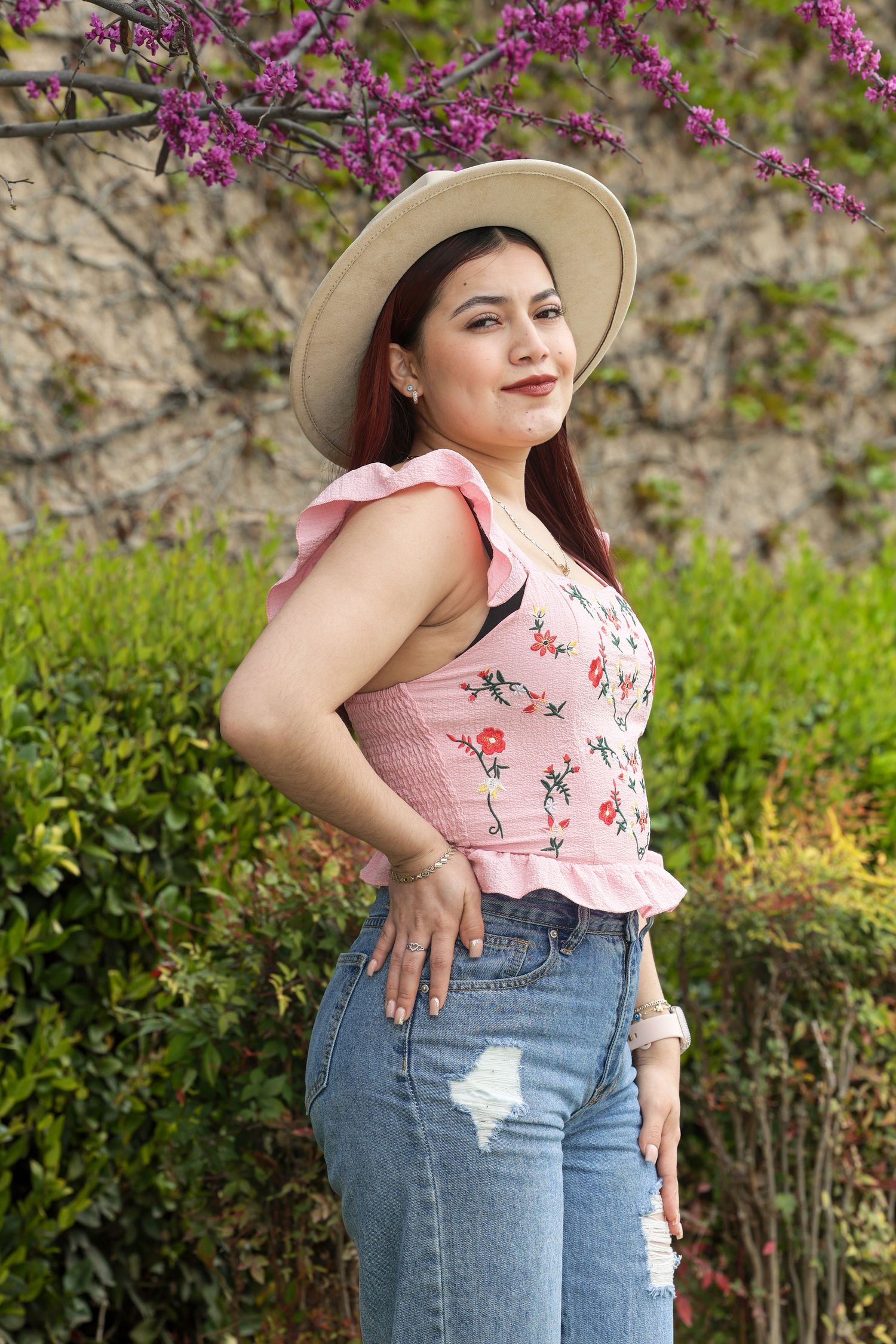 Woman in floral top, jeans and hat posing in front of blooming spring garden.