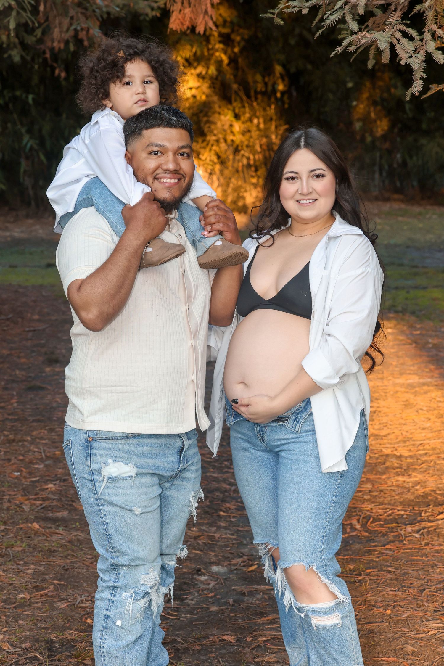 Family maternity portrait with father carrying toddler on shoulders beside pregnant mother in park.