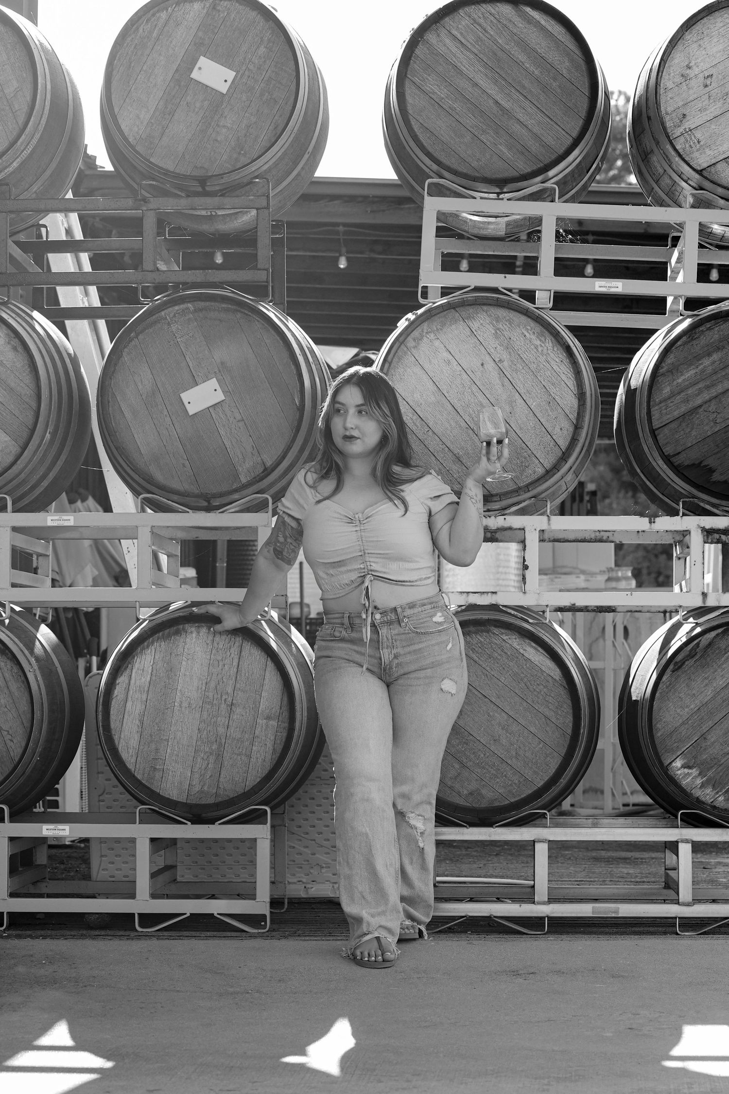 Young woman posing in front of stacked wooden wine barrels in a black and white outdoor winery photo.