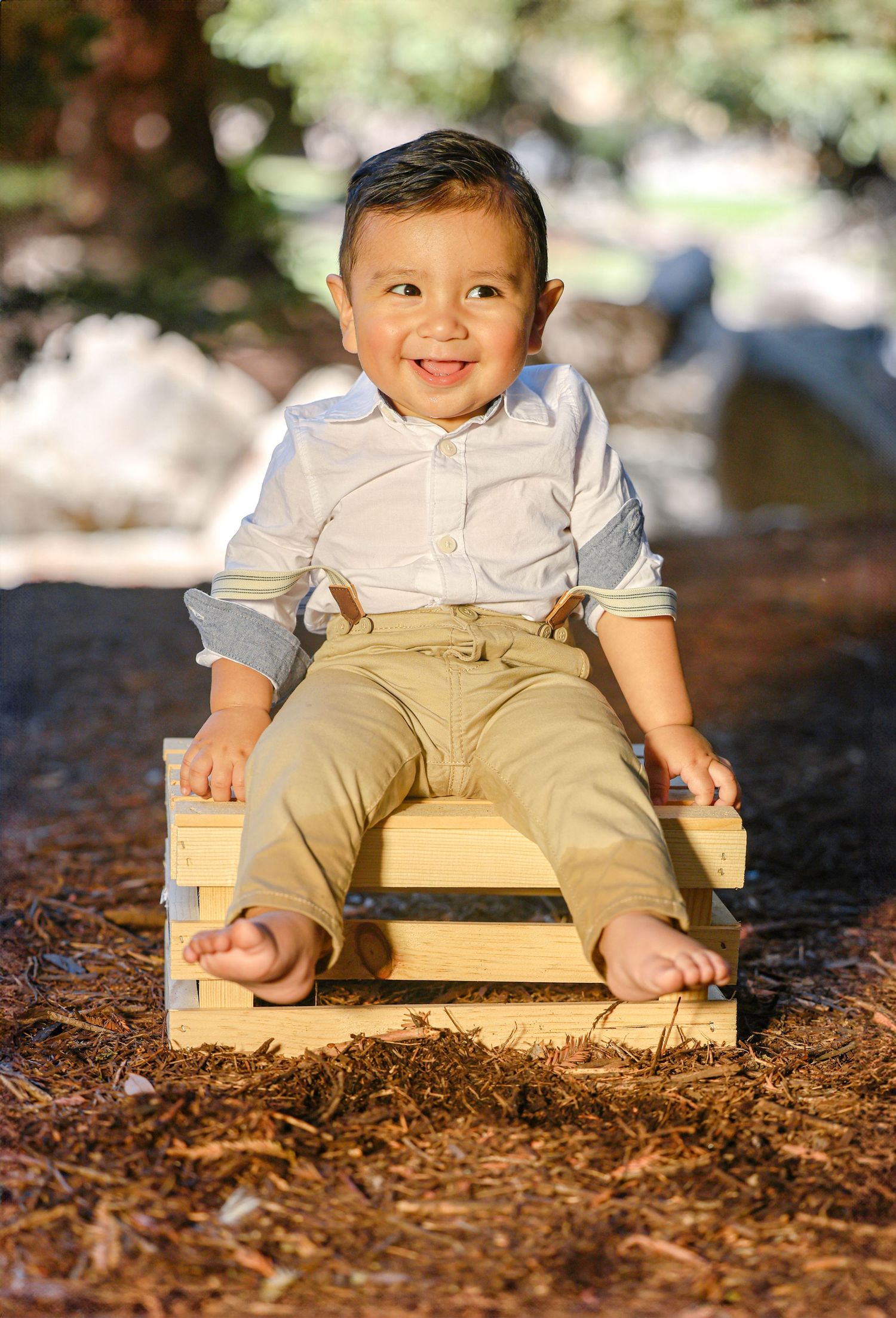 Smiling toddler boy in blue shirt and khakis sits barefoot on wooden crate in sunny outdoor setting.
