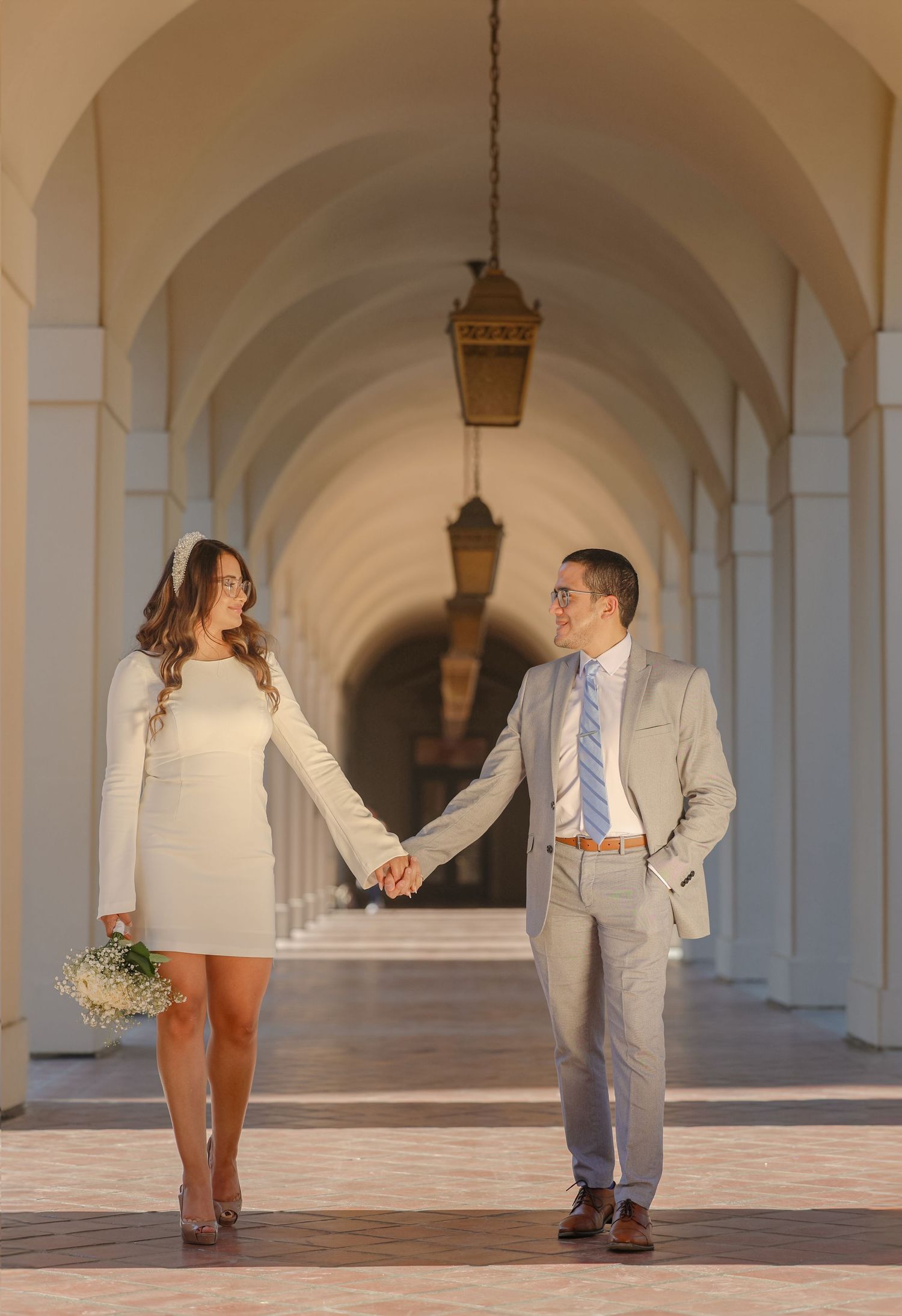 Newlywed couple holding hands walking through elegant arched colonnade in wedding attire.