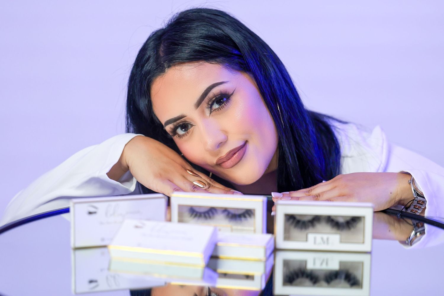 Dark-haired woman resting on reflective surface surrounded by LM branded lash boxes.
