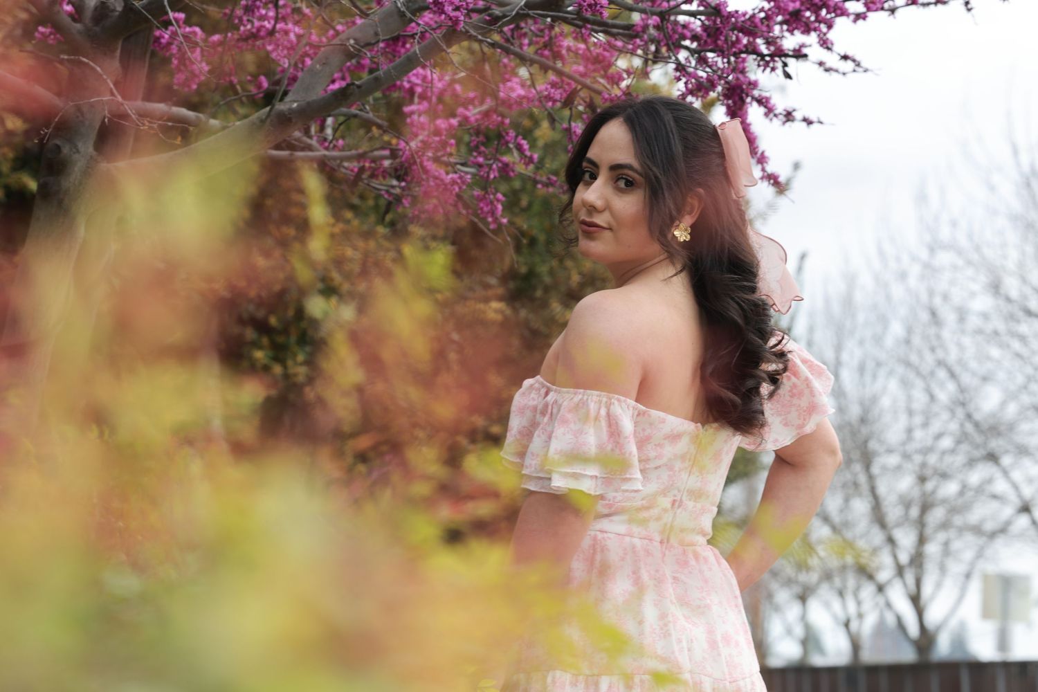 Woman in off-shoulder pink floral dress posing among blooming pink and yellow spring trees outdoors.