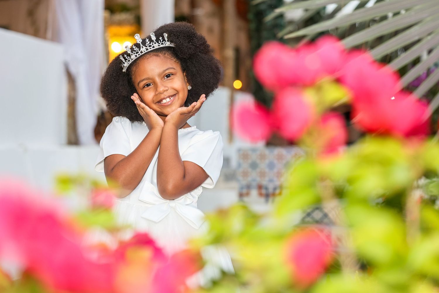 Adorable young girl wearing a tiara and white dress smiling among pink flowers at an outdoor celebration.