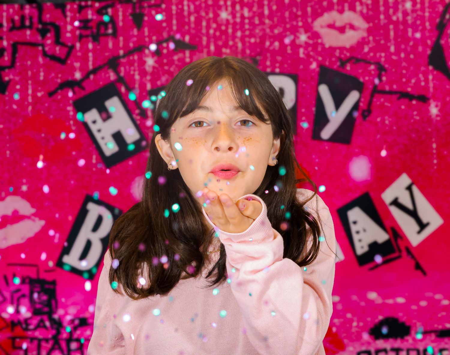 Young girl blowing confetti at a vibrant pink Happy Birthday party celebration backdrop.