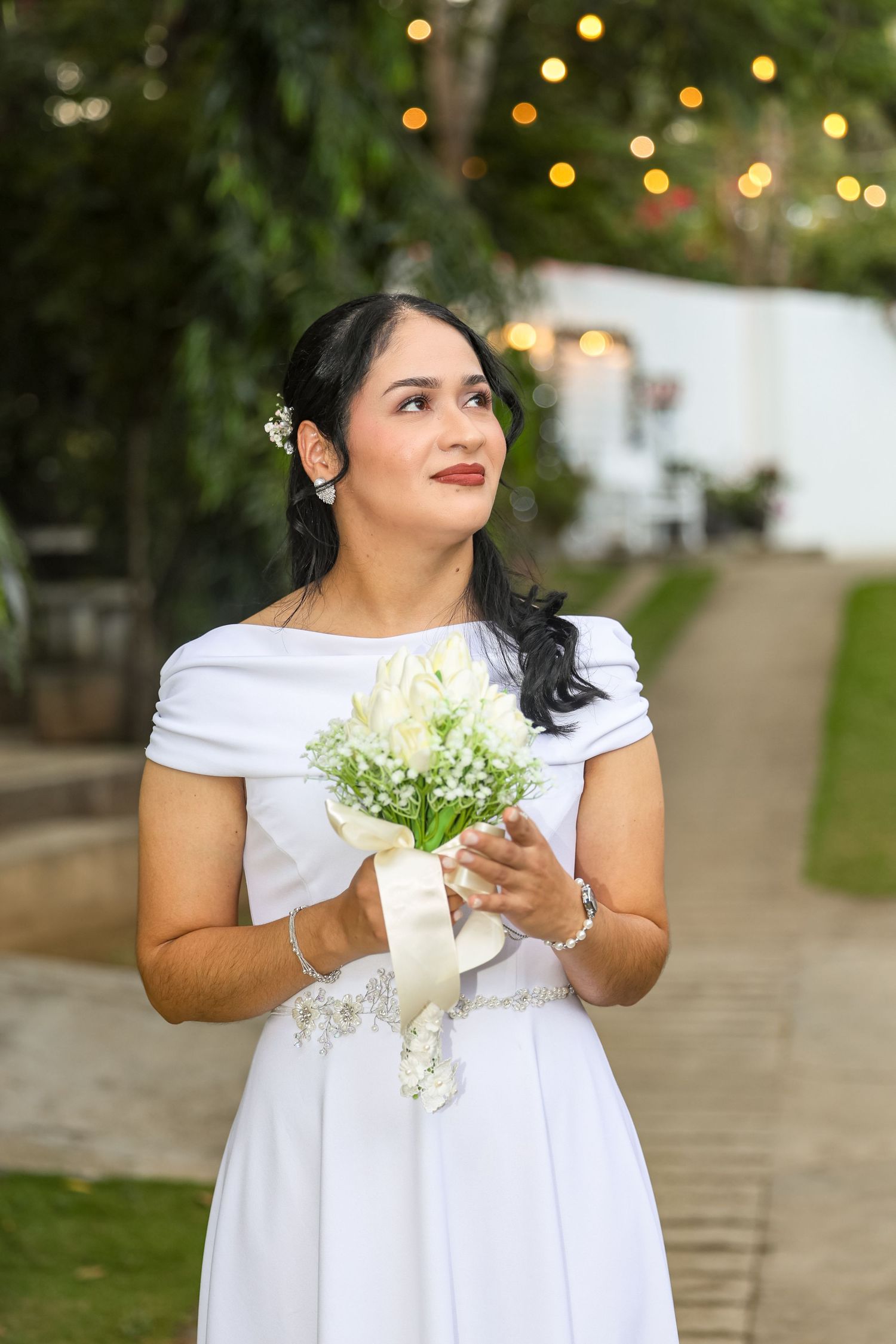 Bride in white off-shoulder dress holds white flower bouquet while gazing upward at outdoor venue.