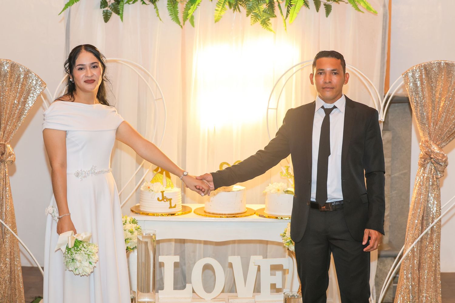 Newlywed couple holds hands beside wedding cake at reception with LOVE sign and floral backdrop.