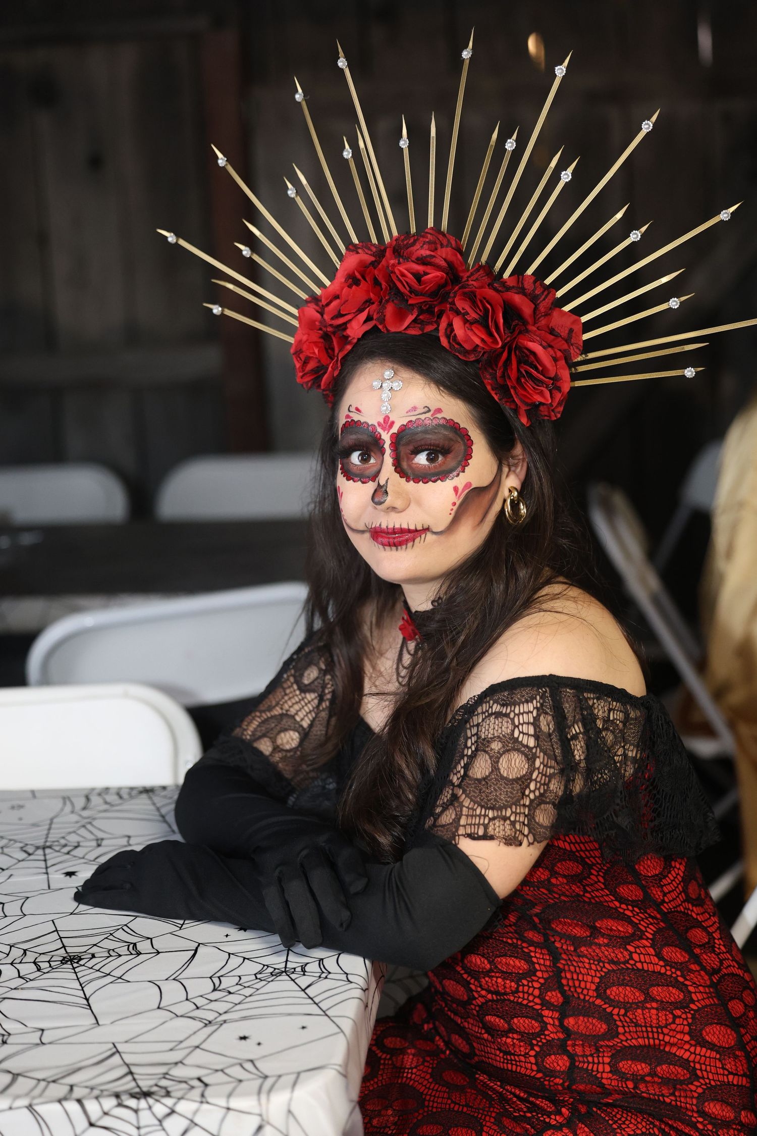 Woman wearing elaborate Day of the Dead sugar skull makeup and red rose crown sits at table.