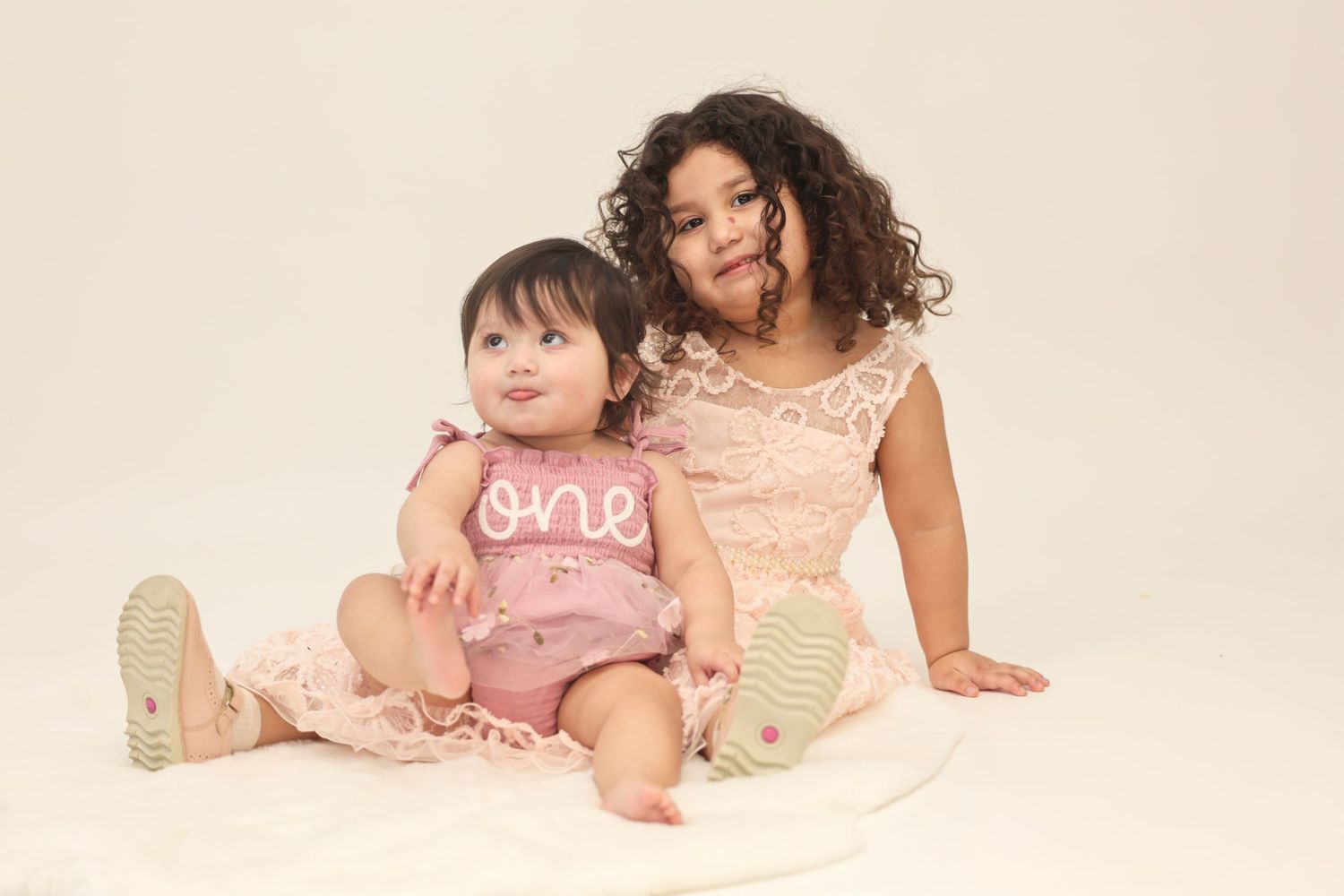 Baby in pink one outfit sitting with older curly-haired sister on white fluffy blanket.