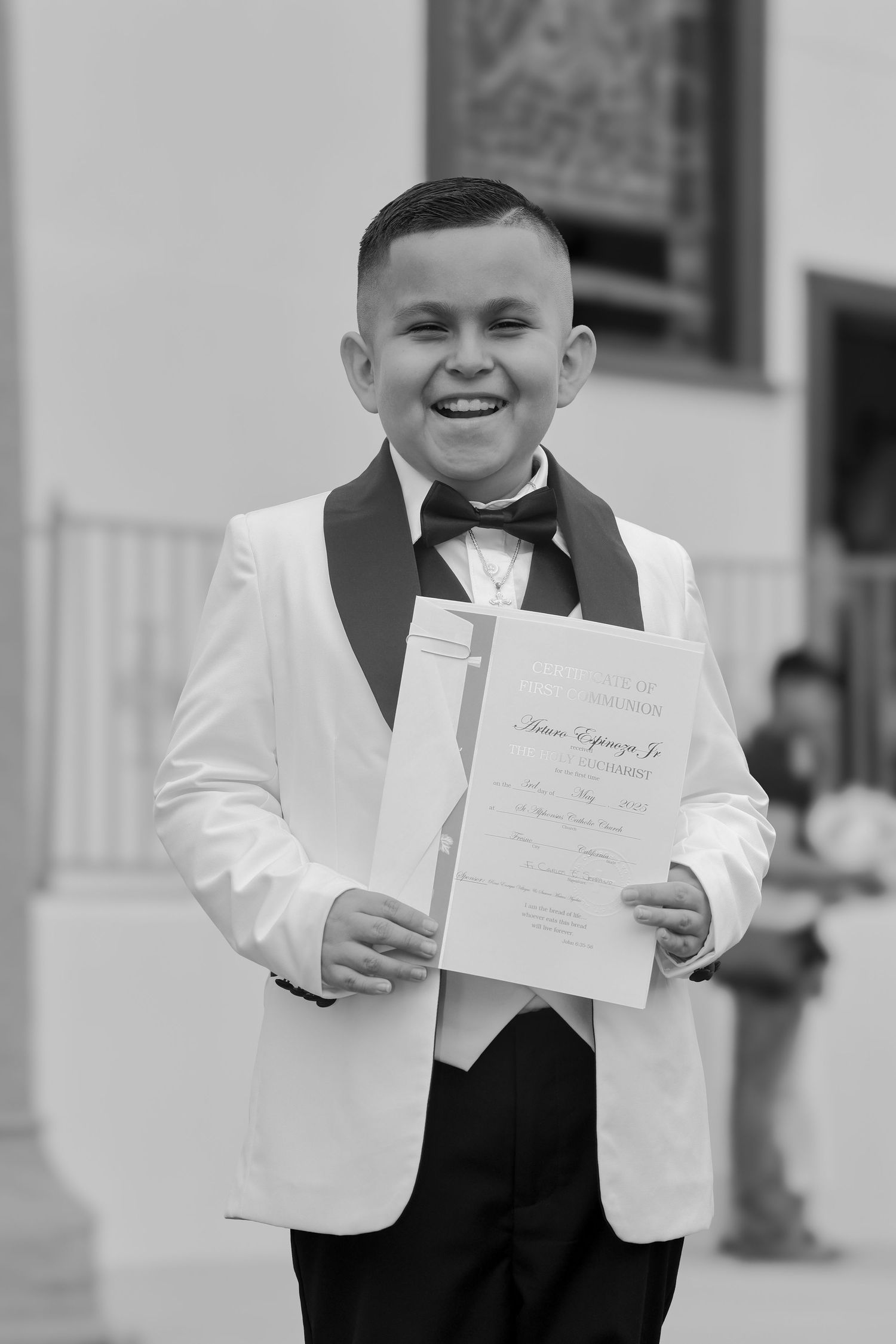 Smiling boy in white tuxedo holding certificate at formal ceremony, black and white photo.