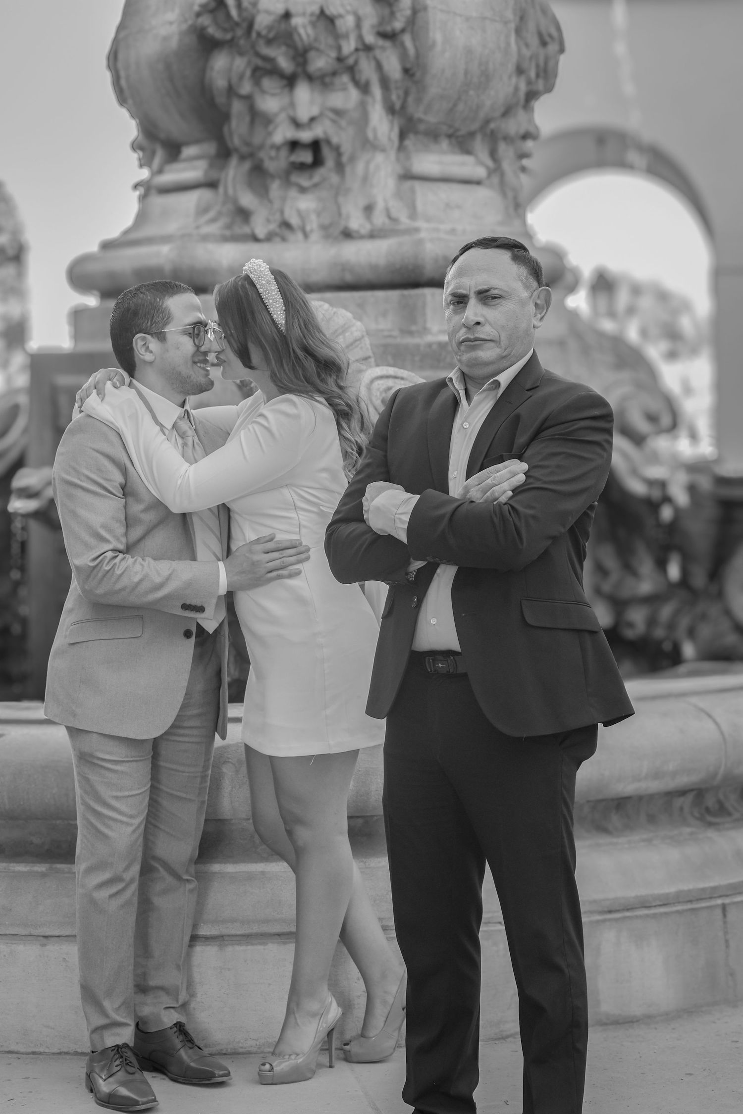 Black and white photo of couple embracing near fountain while man stands with arms crossed.