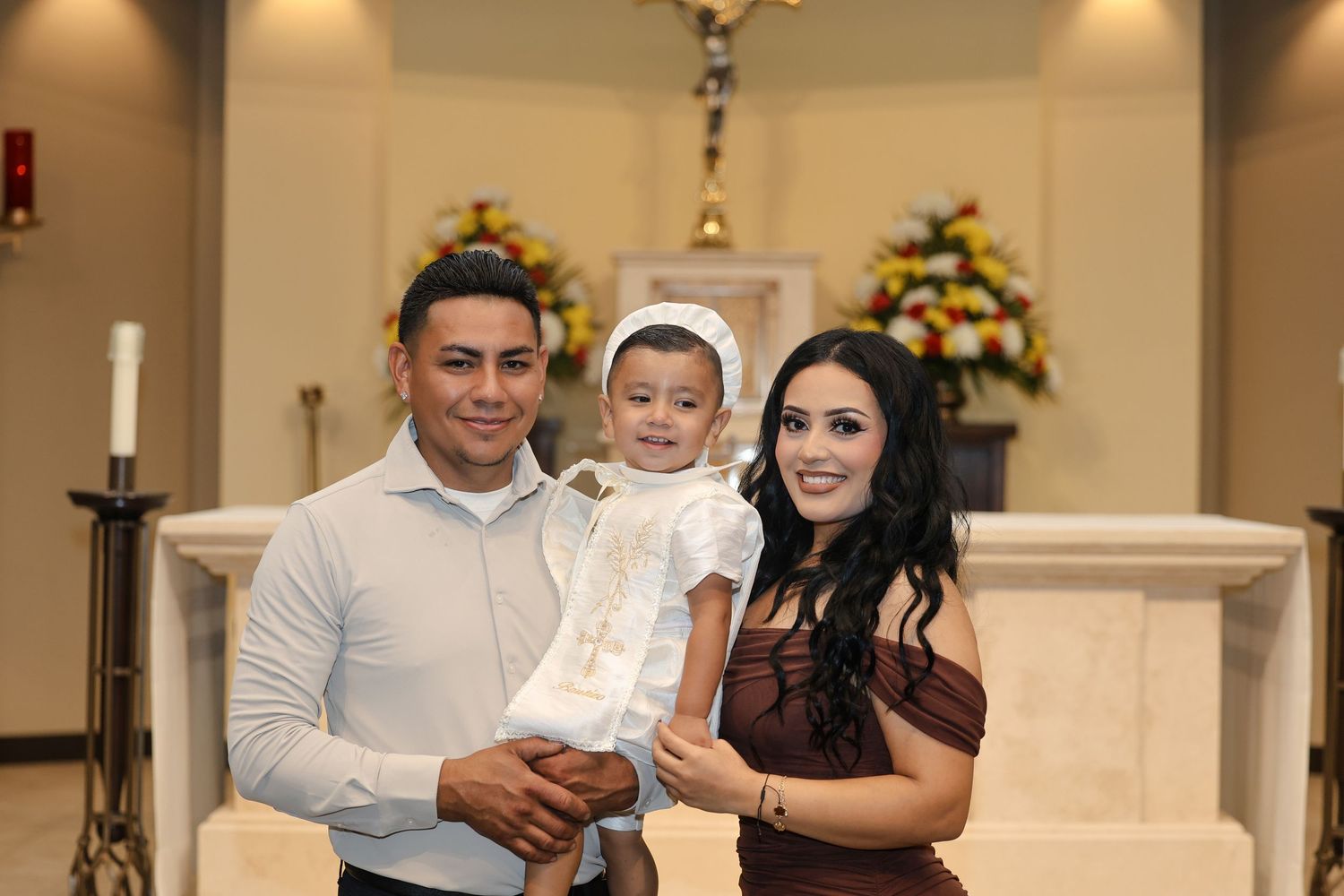 Family of three posing inside church at baptism ceremony, parents holding infant in white christening gown.