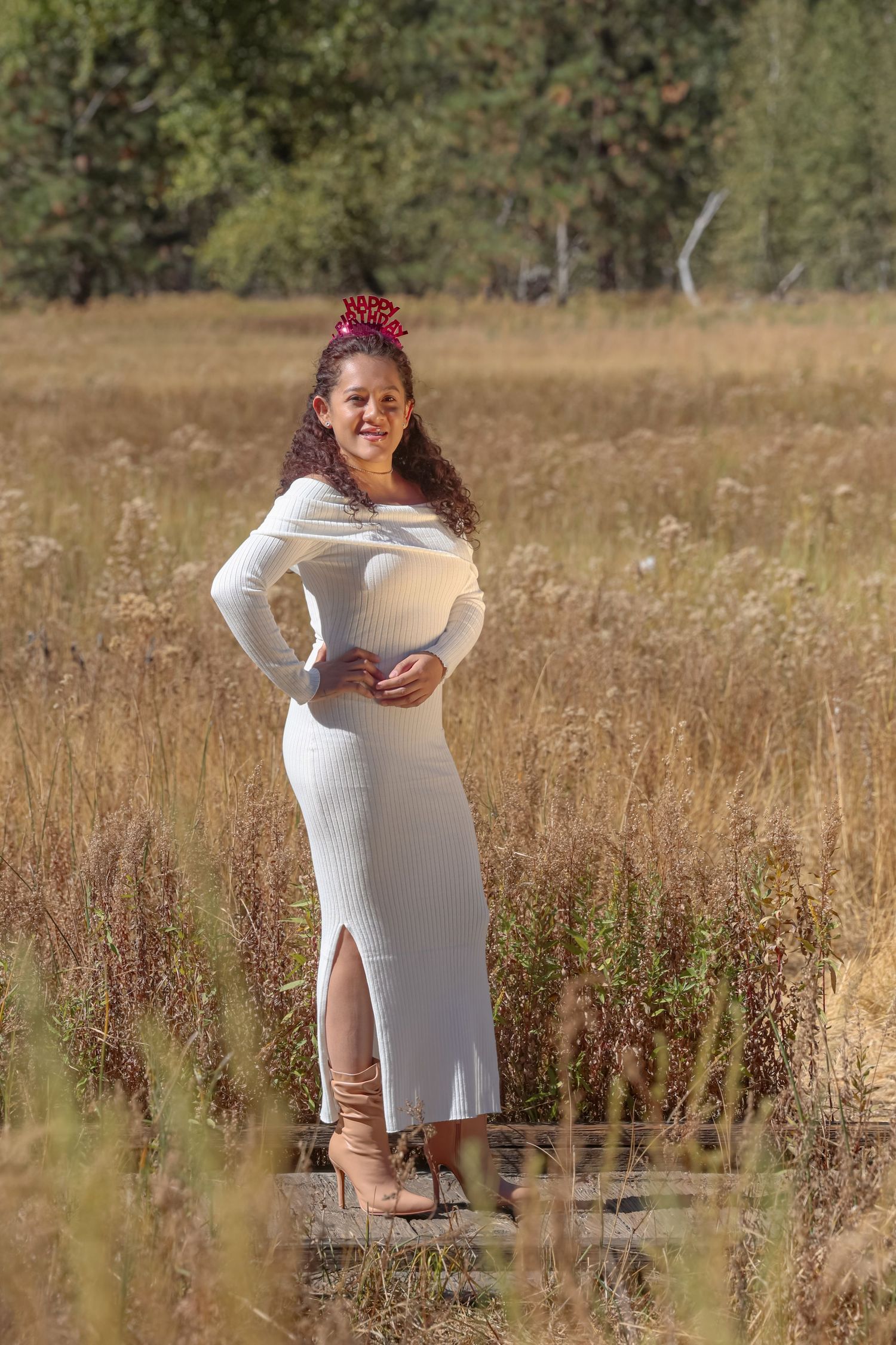 Pregnant woman in white ribbed dress posing in golden autumn field with red bow.