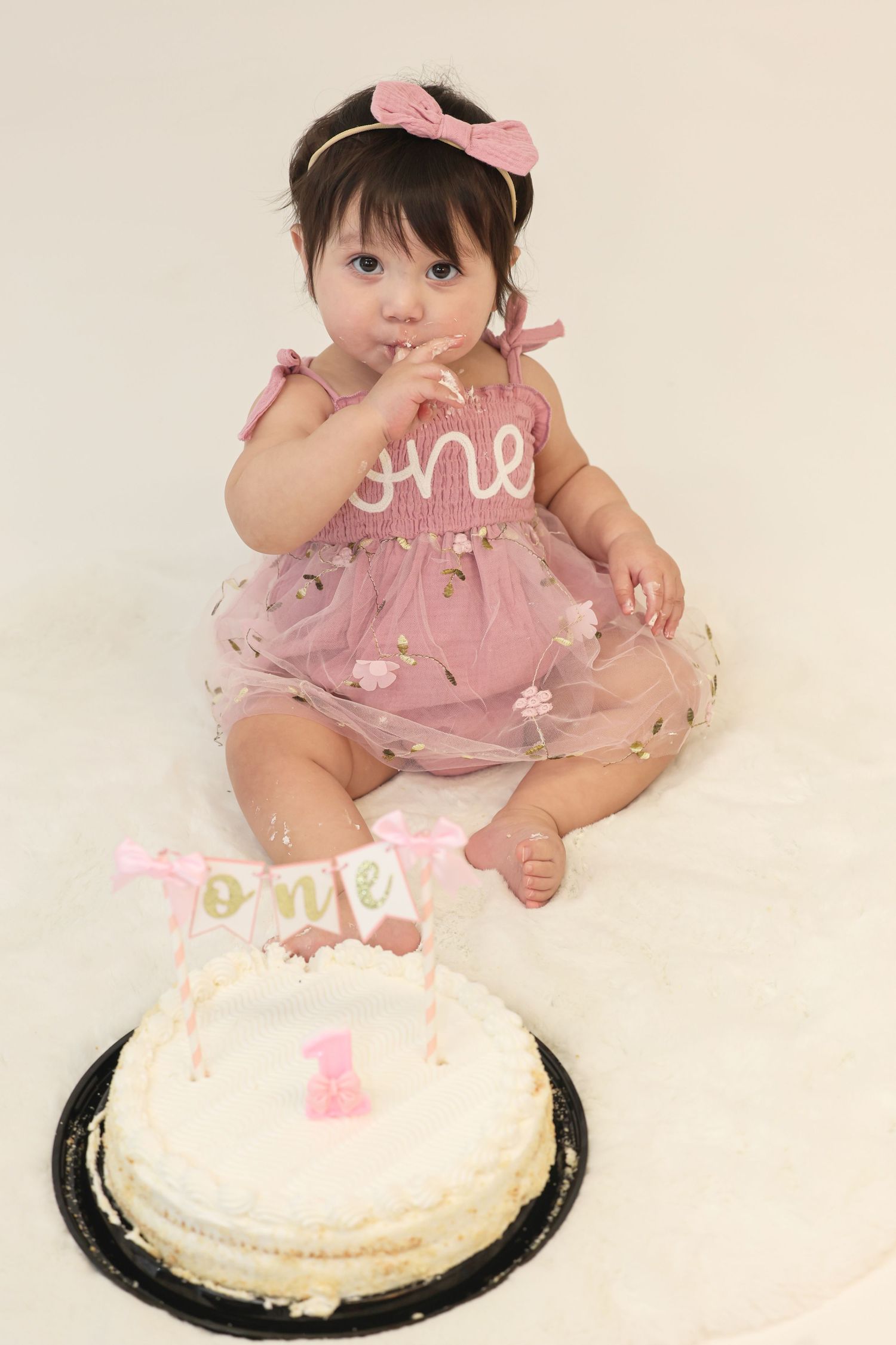 Baby girl in pink tutu dress celebrates first birthday smash cake session with white frosted cake.