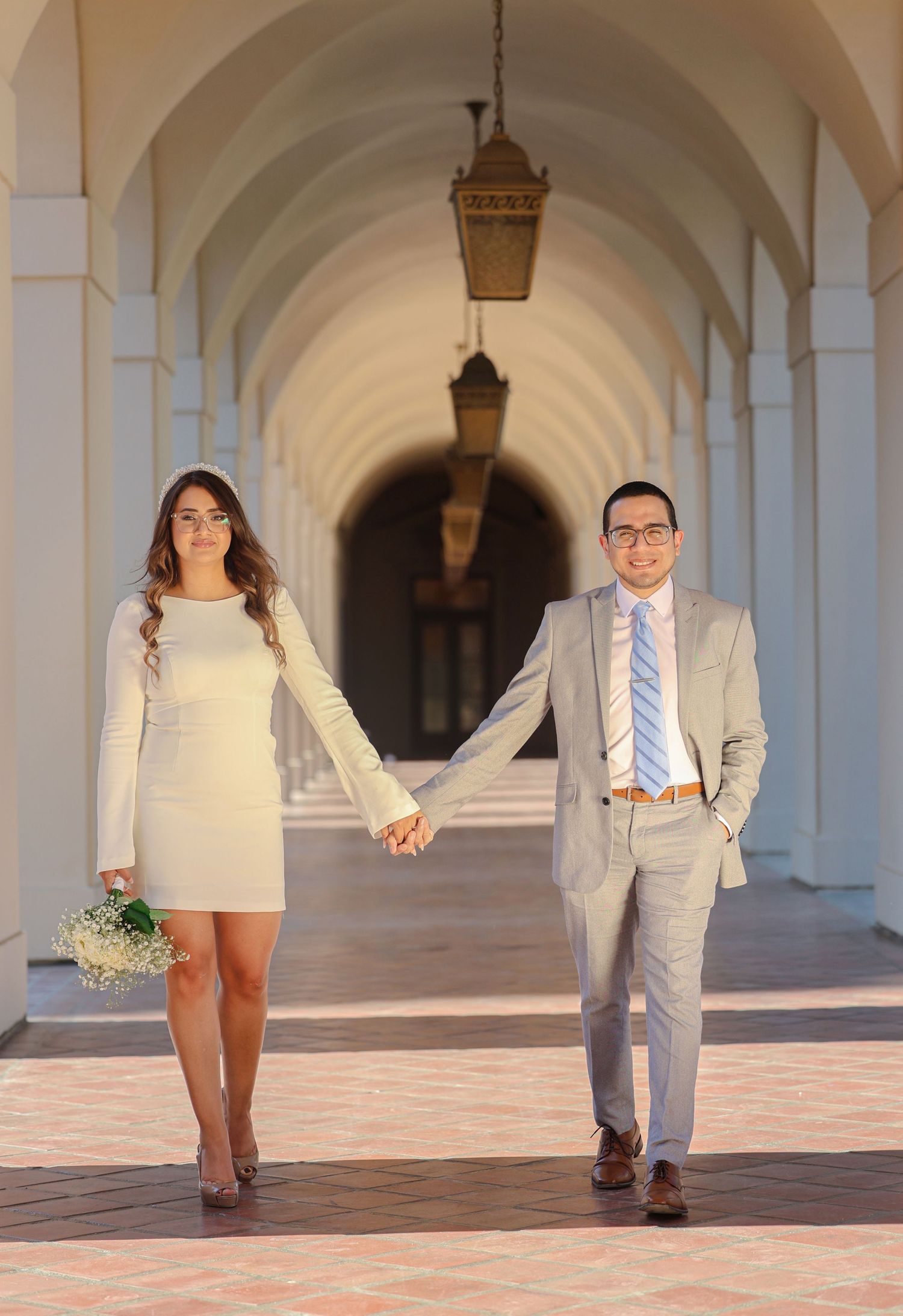 Newlyweds hold hands walking through arched corridor, bride in white dress holding bouquet.