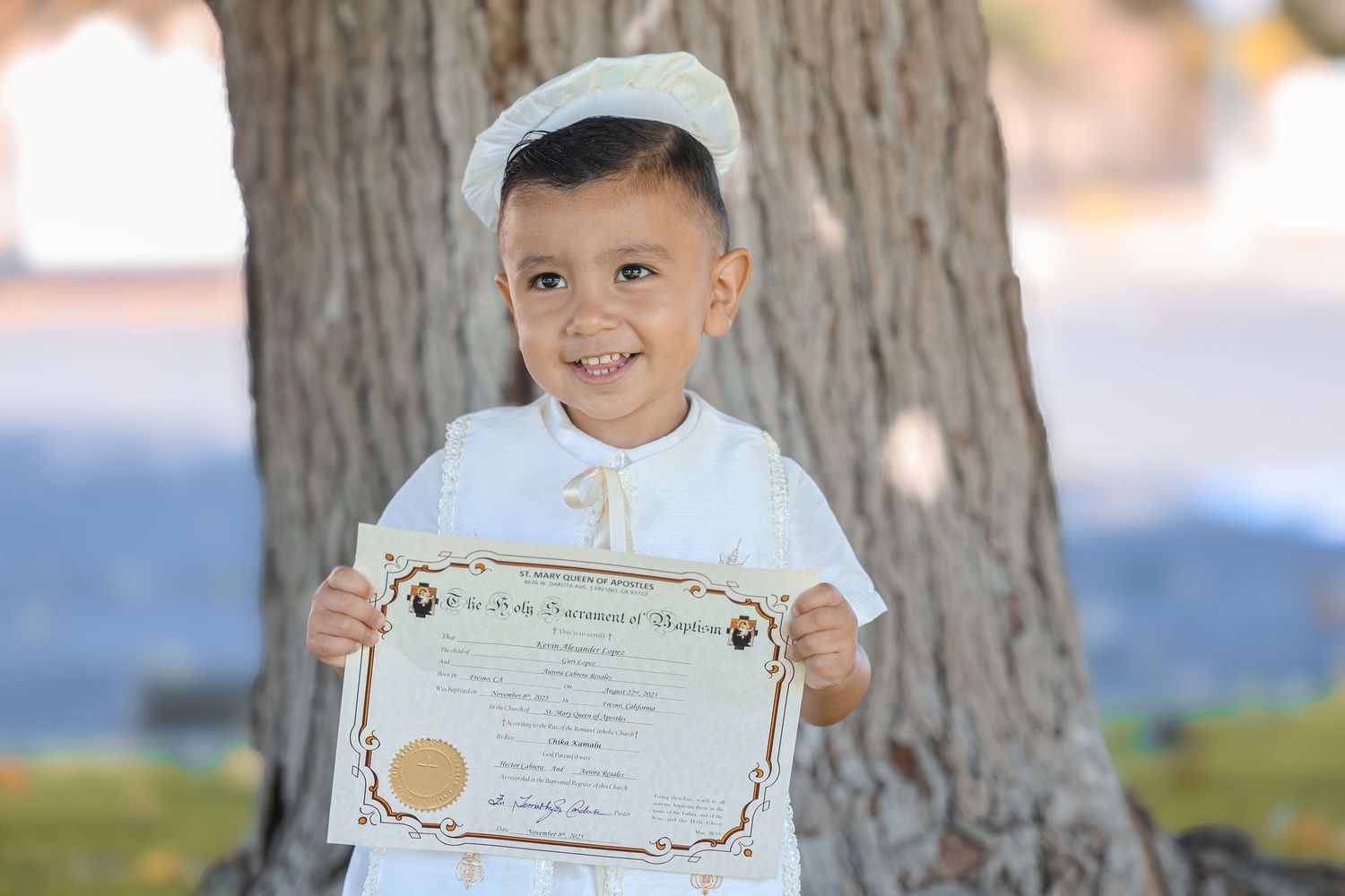 Young boy in white christening outfit proudly holding baptism certificate outdoors near large tree.