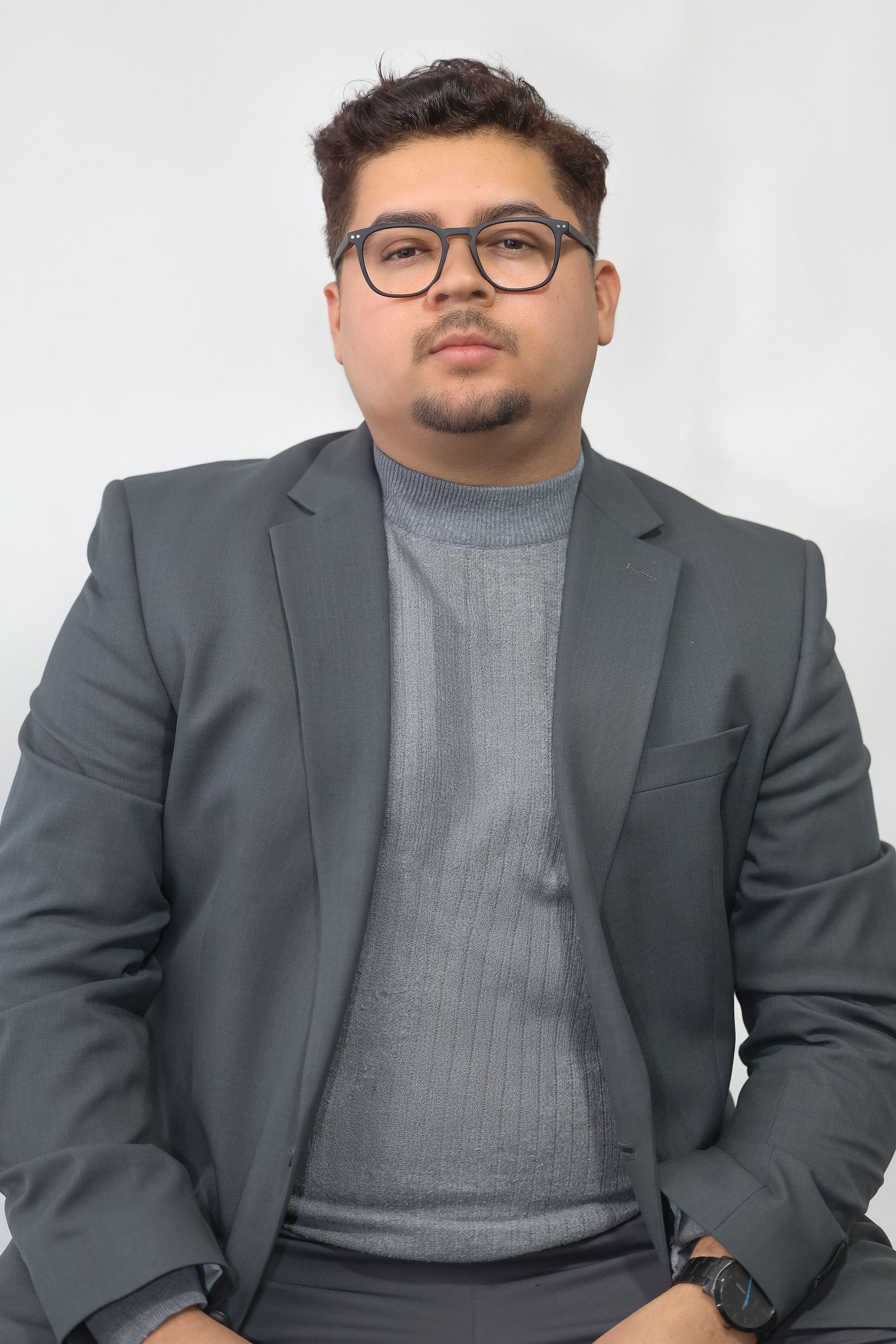 A young man in a gray blazer and glasses sits confidently against a white background.