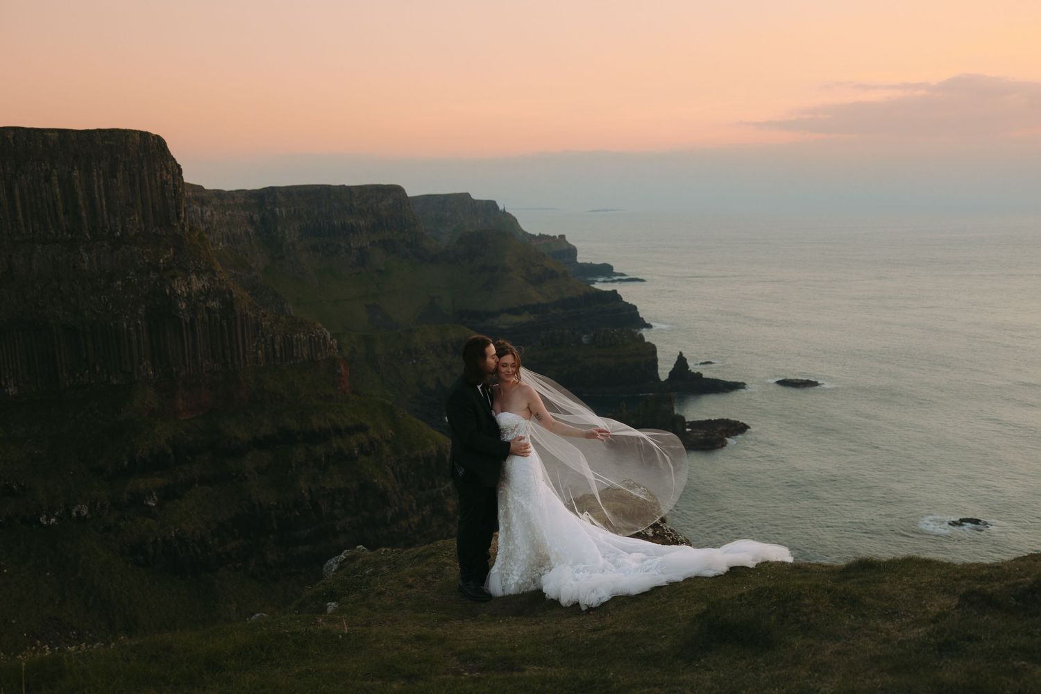 A wedding couple poses next to a helicopter on a grassy hilltop.