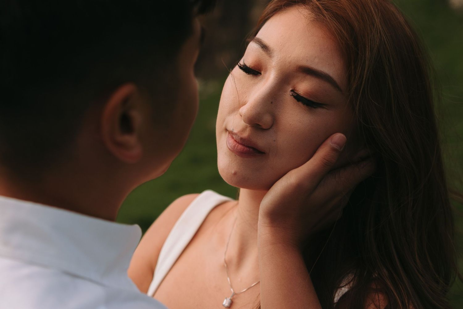 A tender romantic moment between an Asian couple, with a woman closing her eyes as a man gently touches her face outdoors.