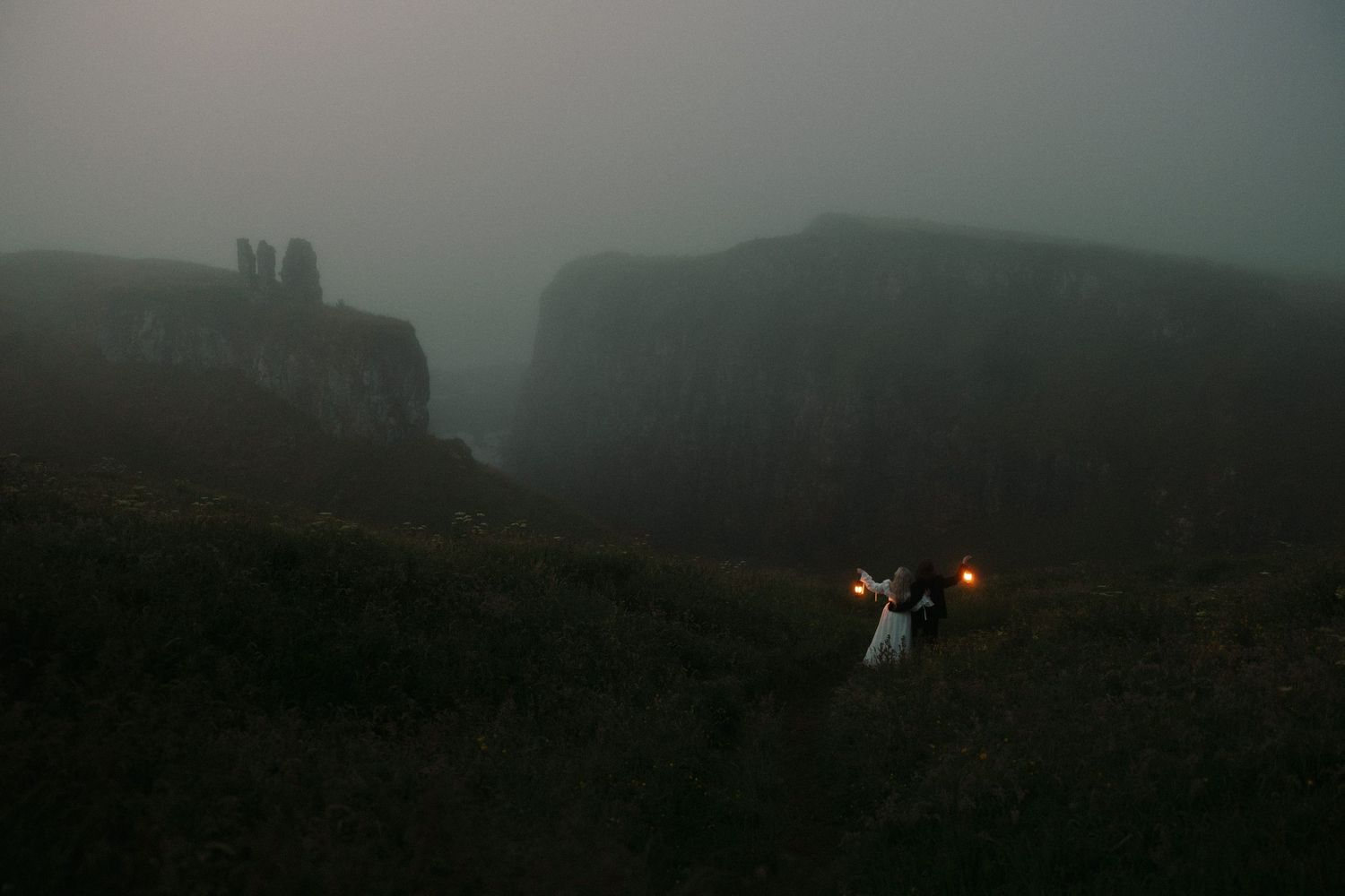 Couple holding torches in misty highland landscape with dramatic foggy cliffs at dusk.
