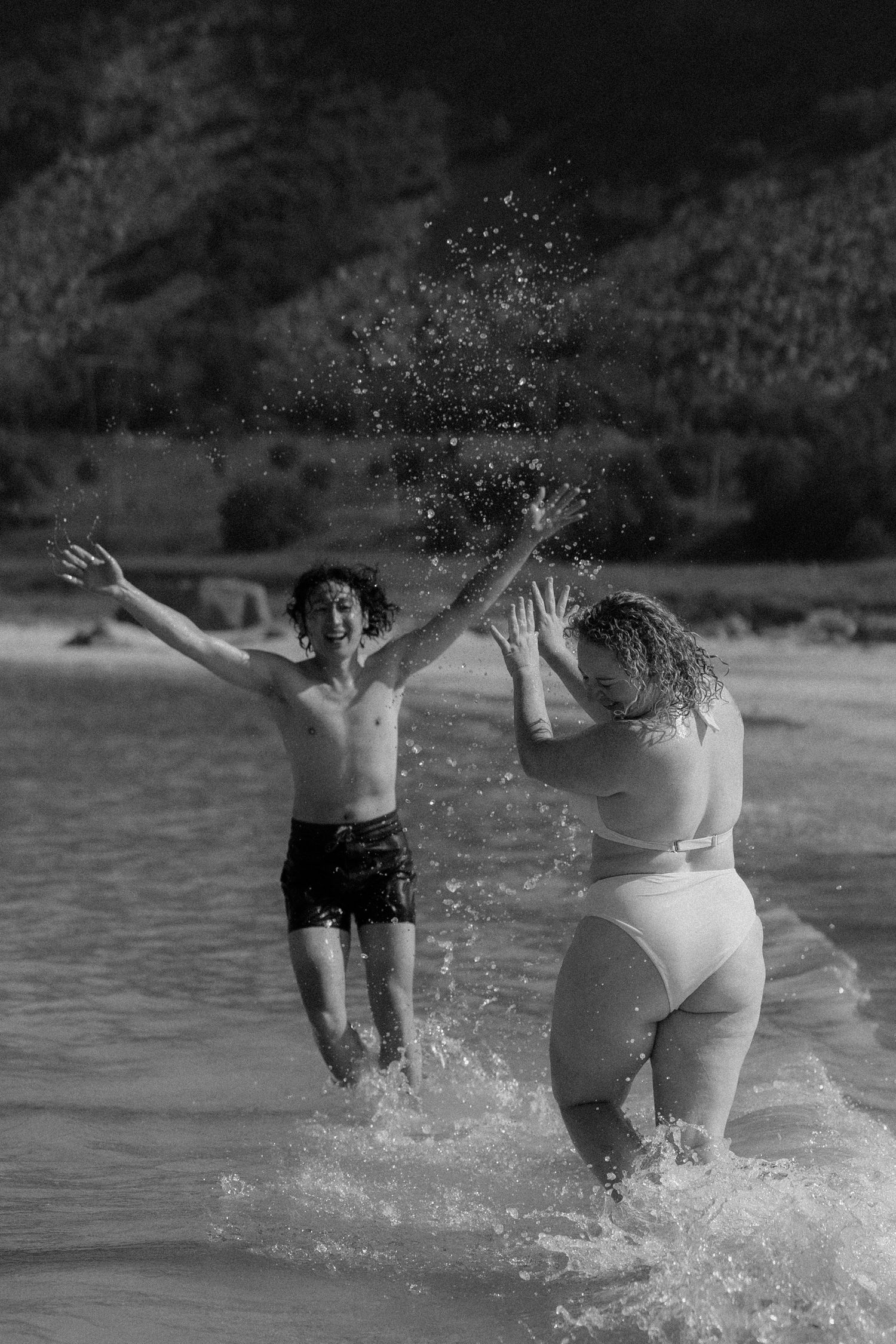 Black and white photo of a woman and boy splashing in ocean waves at a beach, with lush green hills in the background.