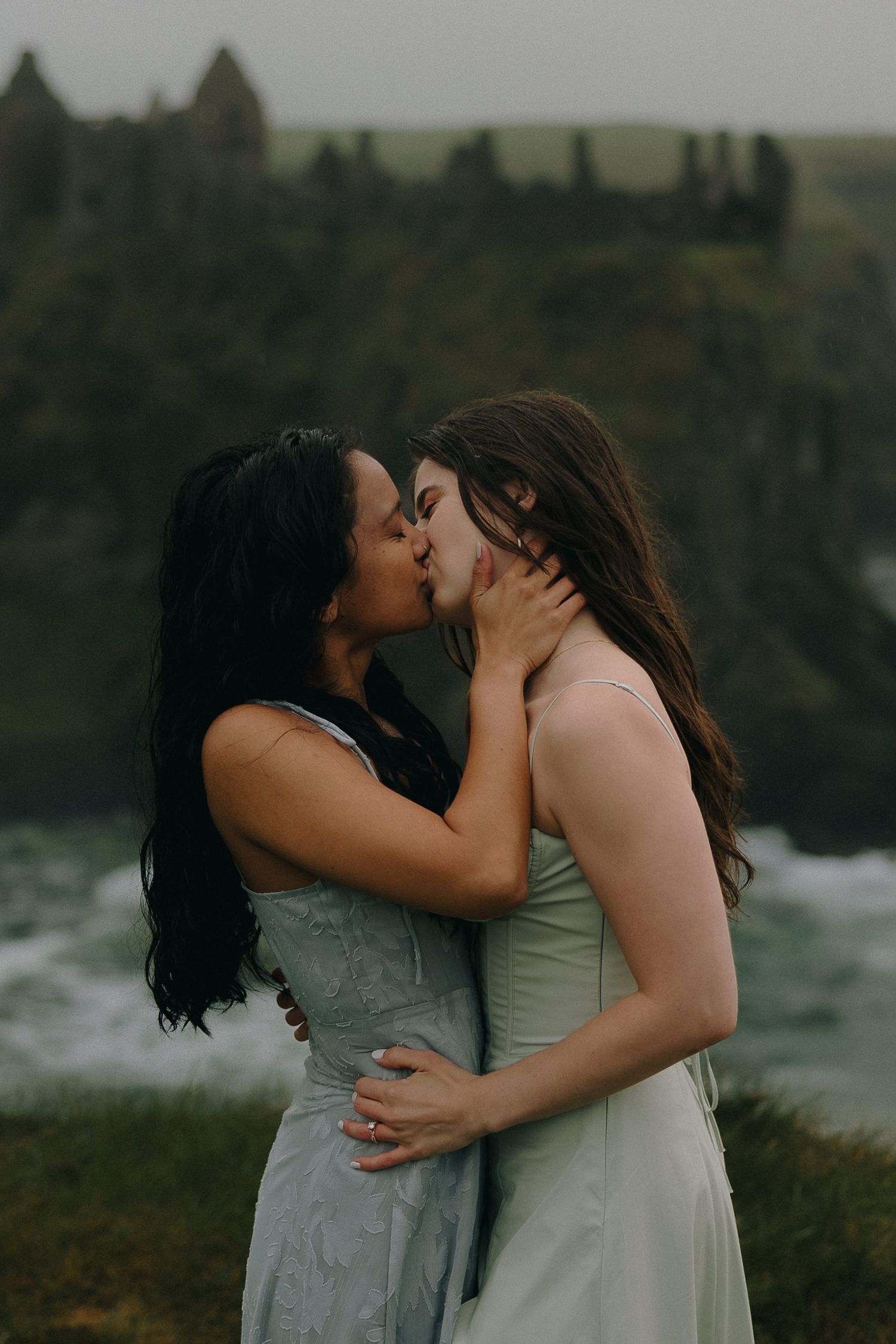 Two brides share a romantic kiss outdoors near a misty coastline with castle ruins in the background.